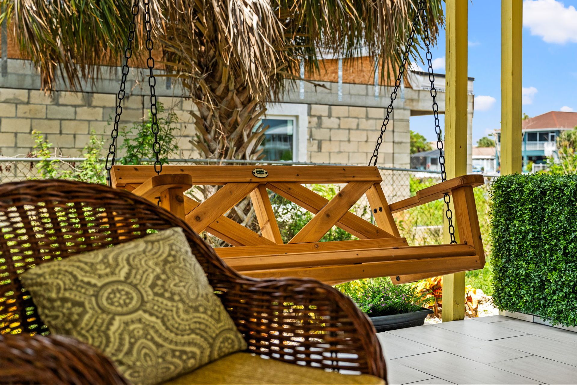 Wicker chair and wooden swing bench on a shaded patio beside a hedge and stone wall.