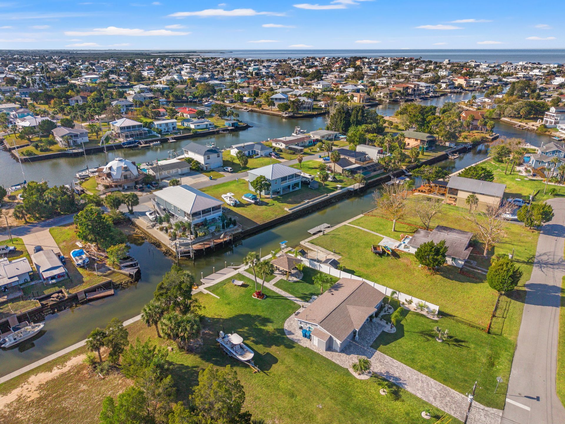 Aerial view of a suburban neighborhood with houses, canals, boats, and a road under a clear blue sky