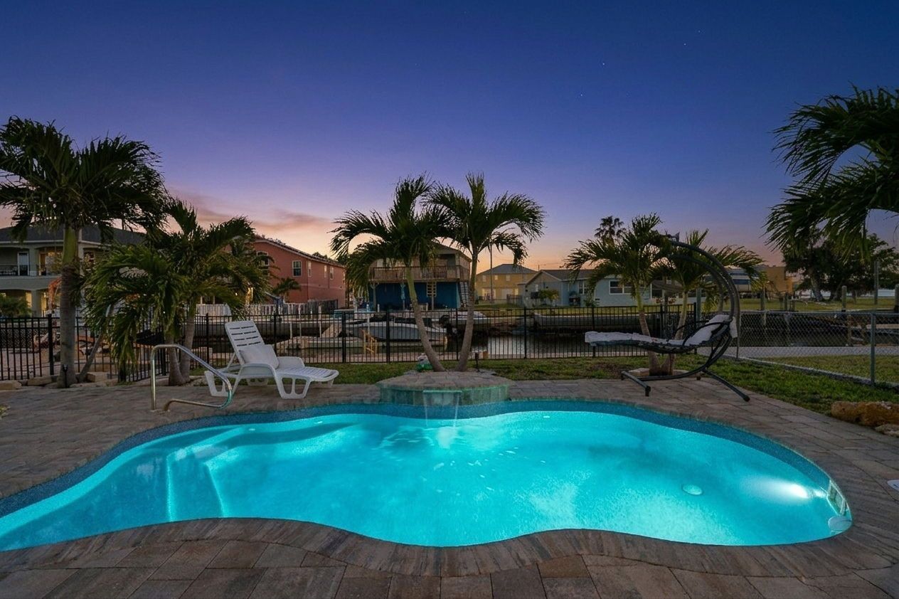 Backyard pool glowing blue at dusk, with palm trees and lounge chairs around it