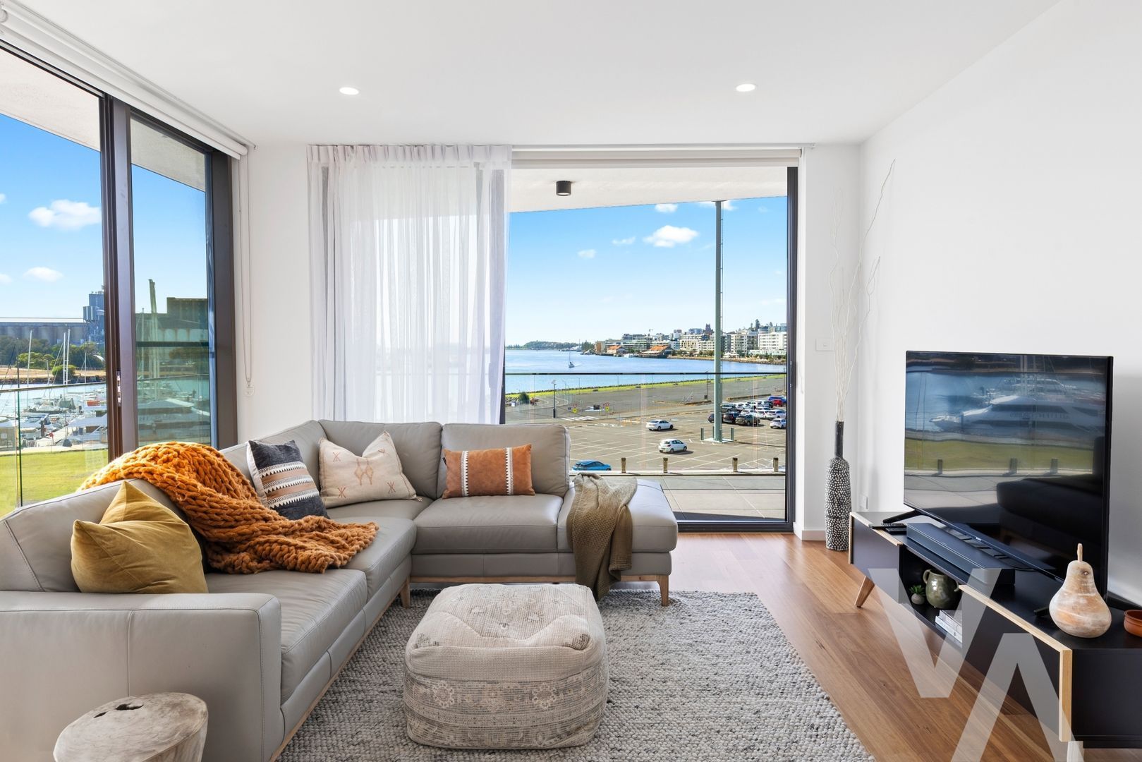 A modern living room with a grey sectional sofa, a furry ottoman, and a large window looking out onto a coastal cityscape.