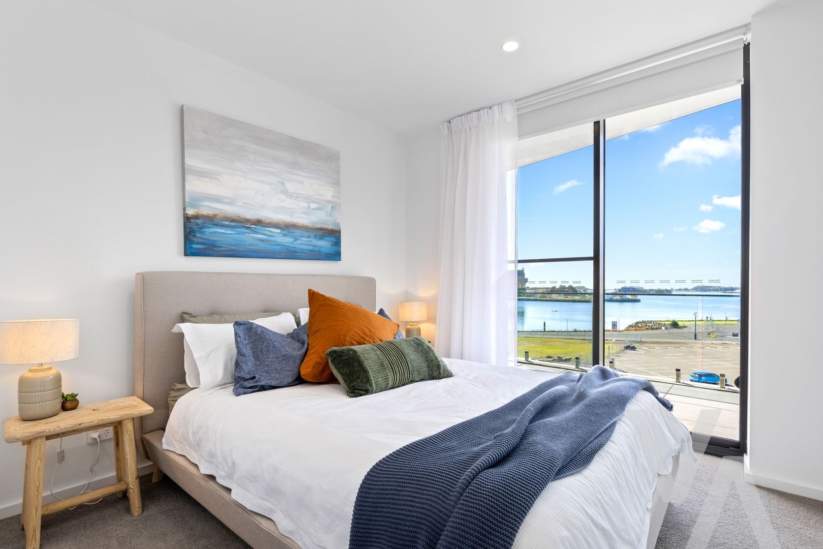 A modern bedroom featuring a bed with white bedding, throw pillows, and a blue blanket, beside a large window with a view.