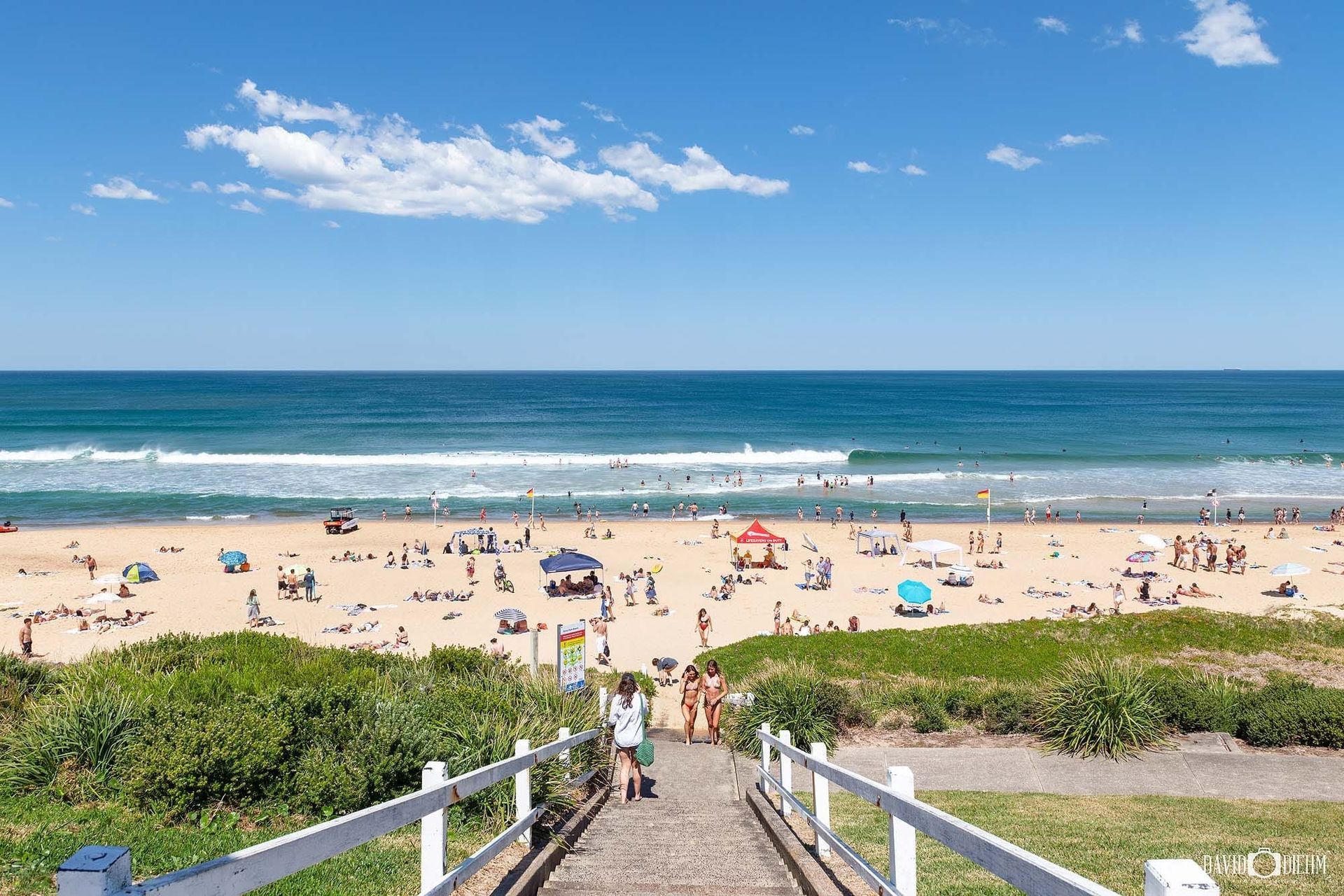 Beach scene with people, blue ocean, sunny sky, and a path leading down. Newcastle NSW