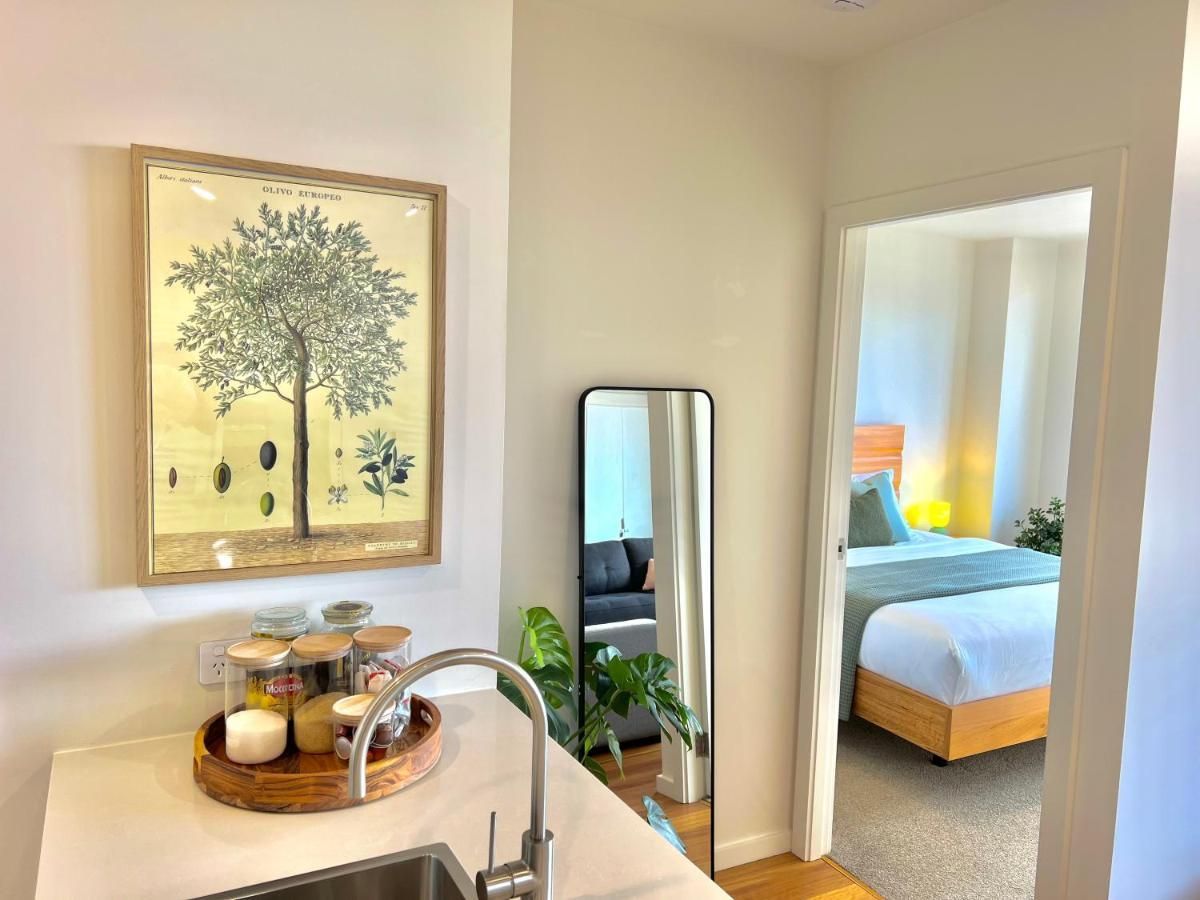 A kitchen sink area with a wooden tray of jars, a framed botanical print, and a mirror looking into a bedroom.