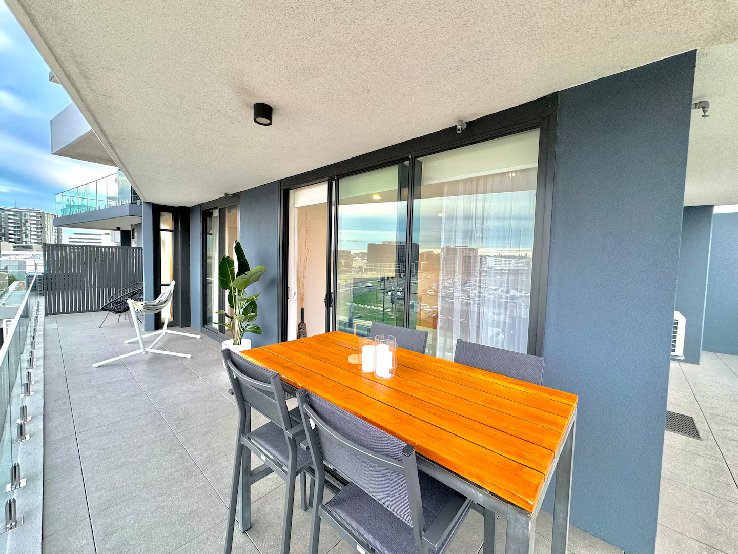 Modern outdoor balcony with a wooden dining table, gray chairs, and a potted plant against a dark blue wall.