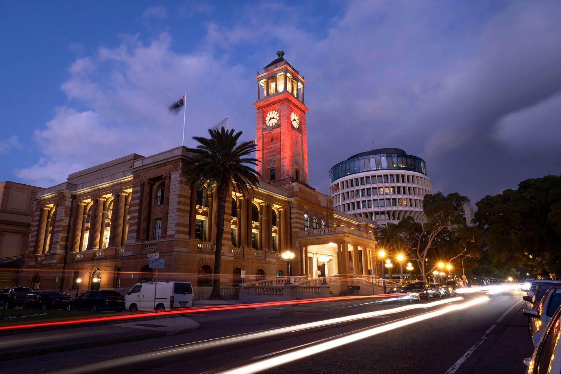 Lit-up clock tower with building, palm tree and modern structure at dusk. Streaks of light from passing cars. Newcastle NSW