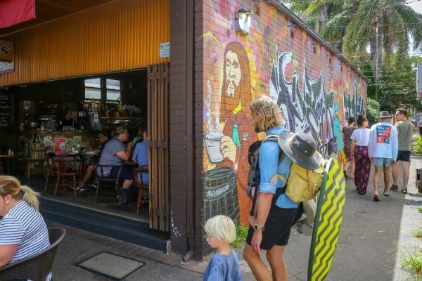 People walking past a cafe with mural art on the wall. A person carries a surfboard. Newcastle NSW