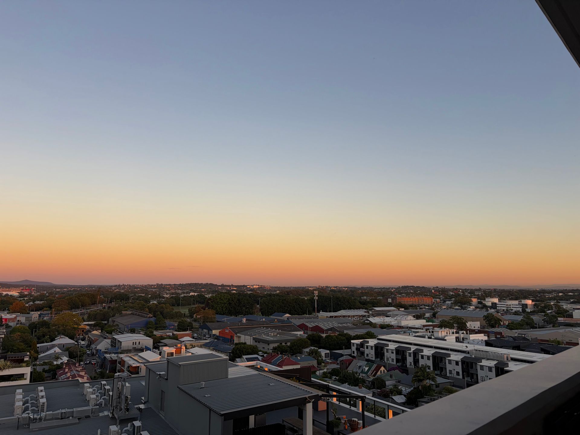Sunset over a cityscape with residential rooftops and industrial buildings under a gradient sky from blue to orange.