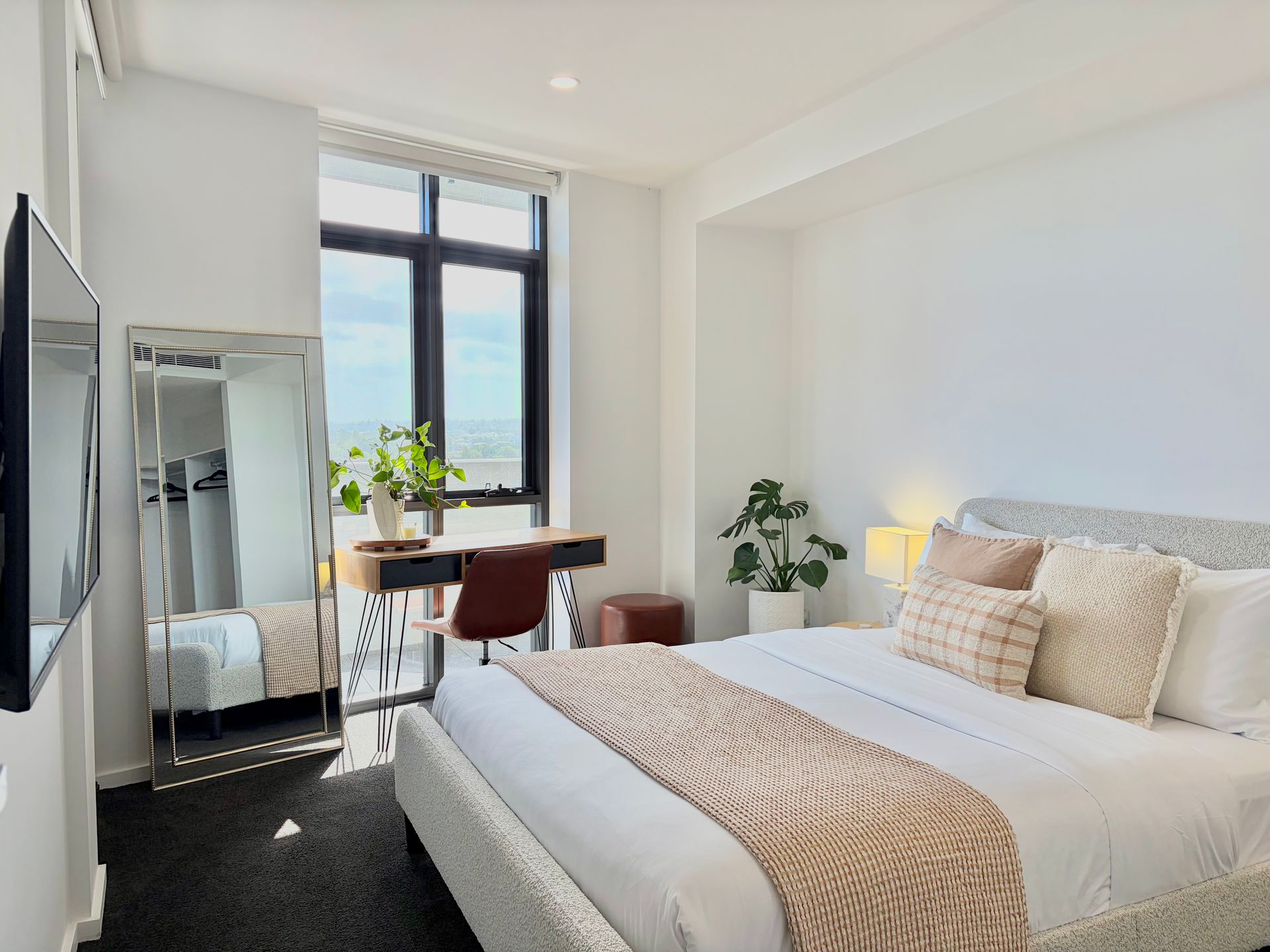 A modern, neutral-toned bedroom with a queen bed, a desk by the window, a large floor mirror, and a television.