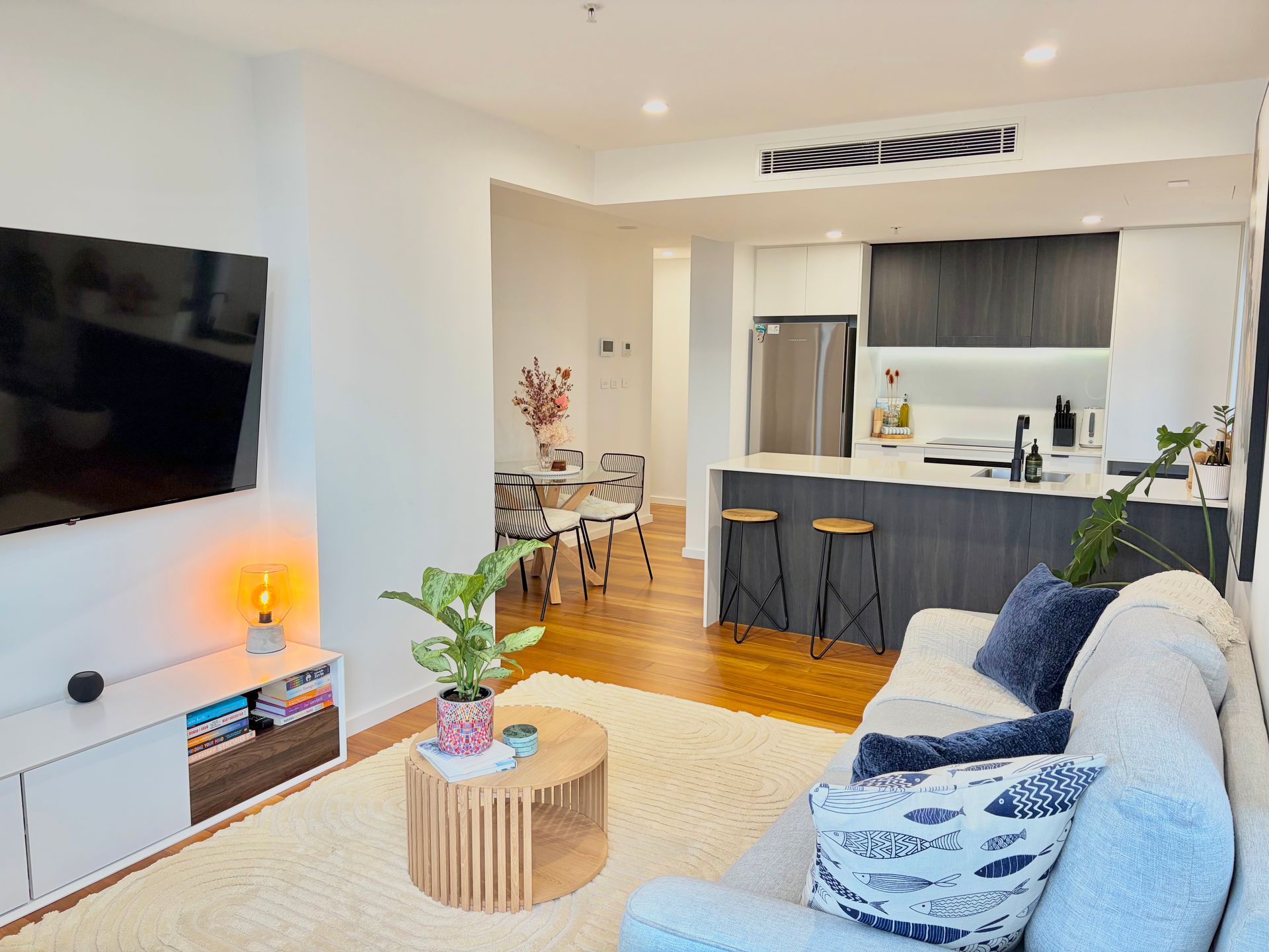 A modern apartment living room with a gray sofa, wood coffee table, media console, and an open-plan kitchen with stools.