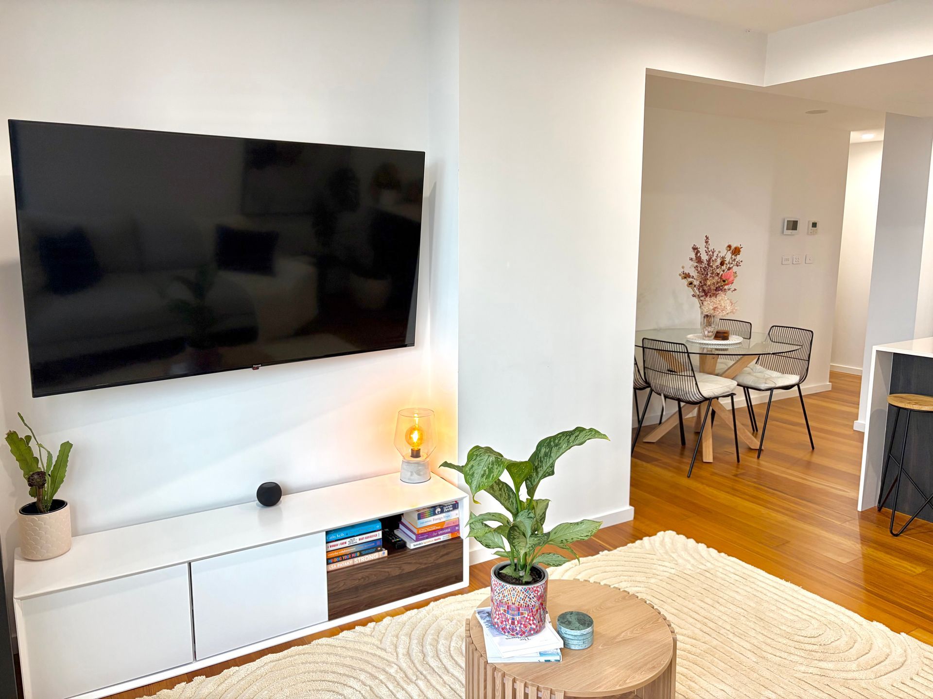 A living room with a wall-mounted TV, white media console, wood coffee table with a plant, and a dining area in the back.