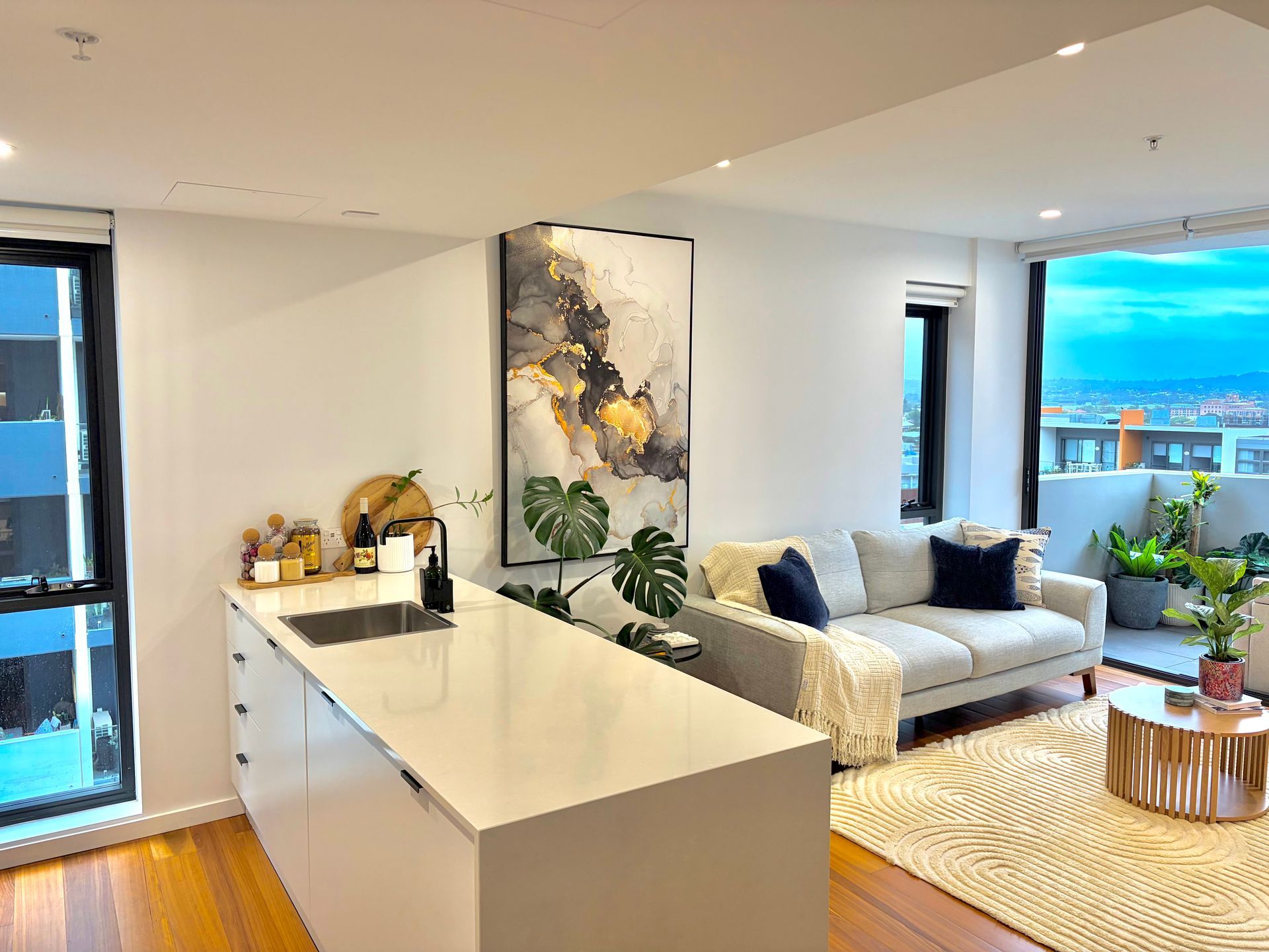 A modern apartment featuring a kitchen island, a gray sofa, abstract art, and a balcony with plants at dusk.