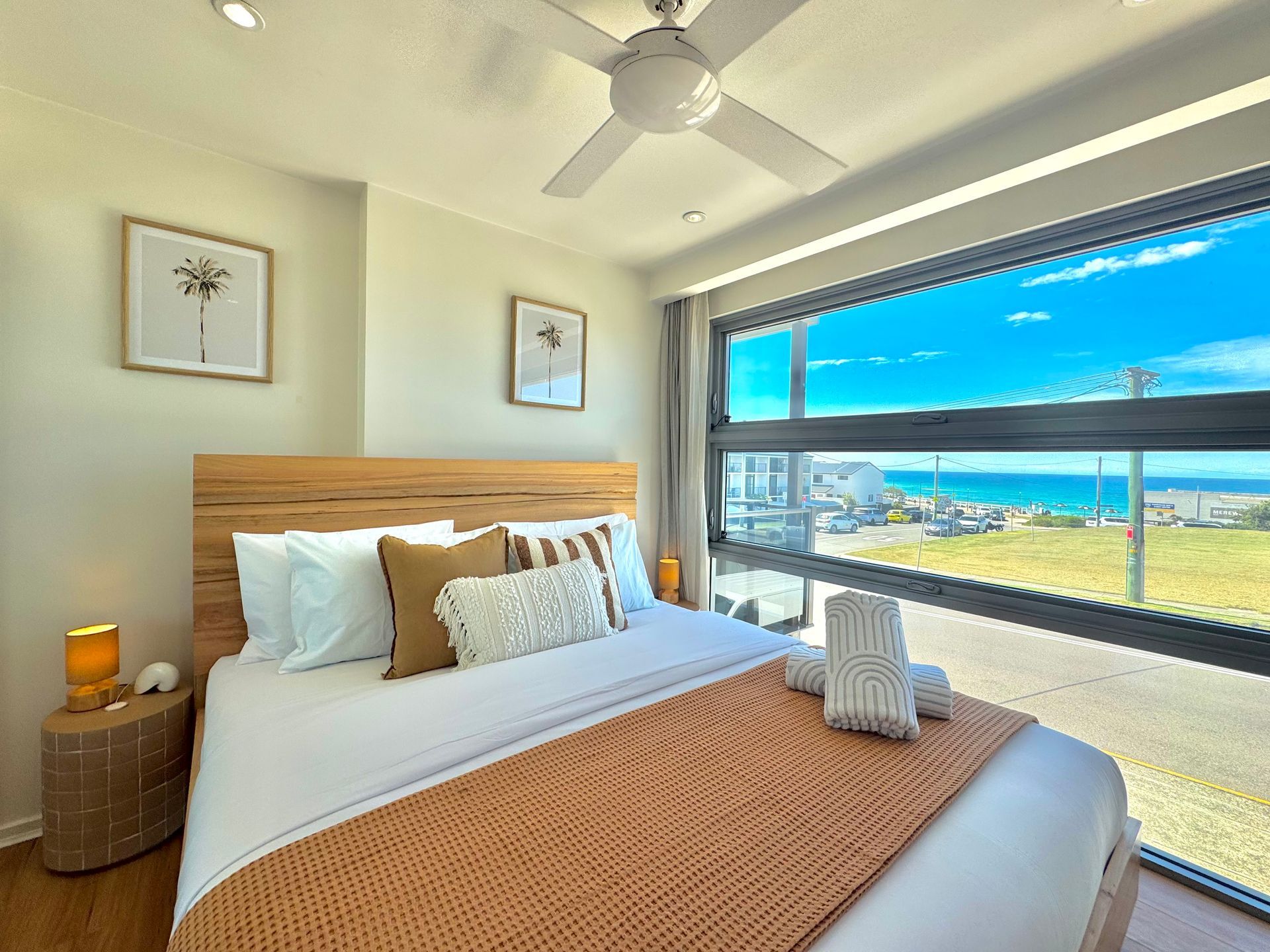 A bright bedroom with a queen bed, wooden headboard, textured orange throw, and large window view of the ocean.