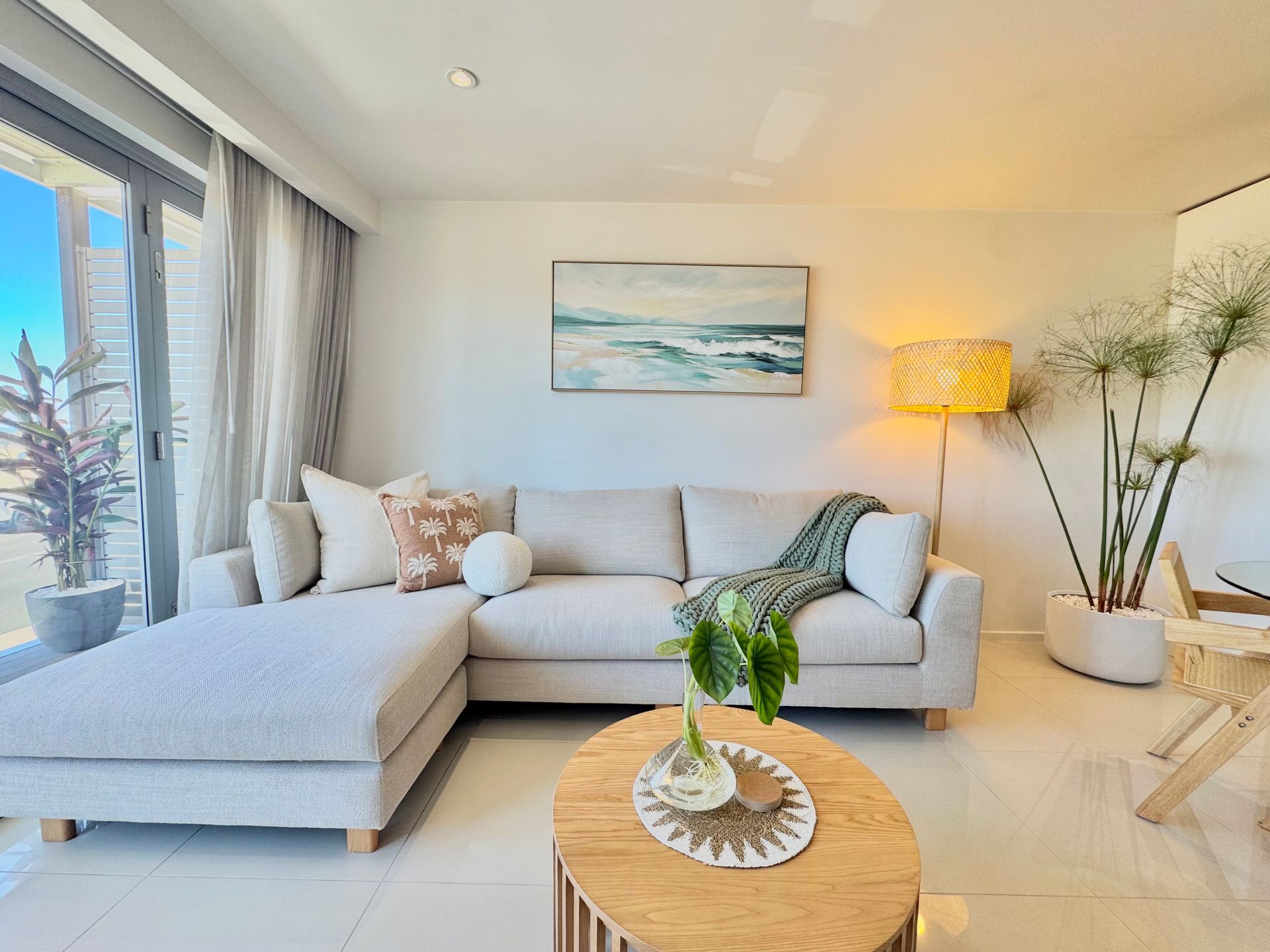 A bright living room features a beige sectional sofa, a round wooden coffee table, a tall floor lamp, and a large plant.