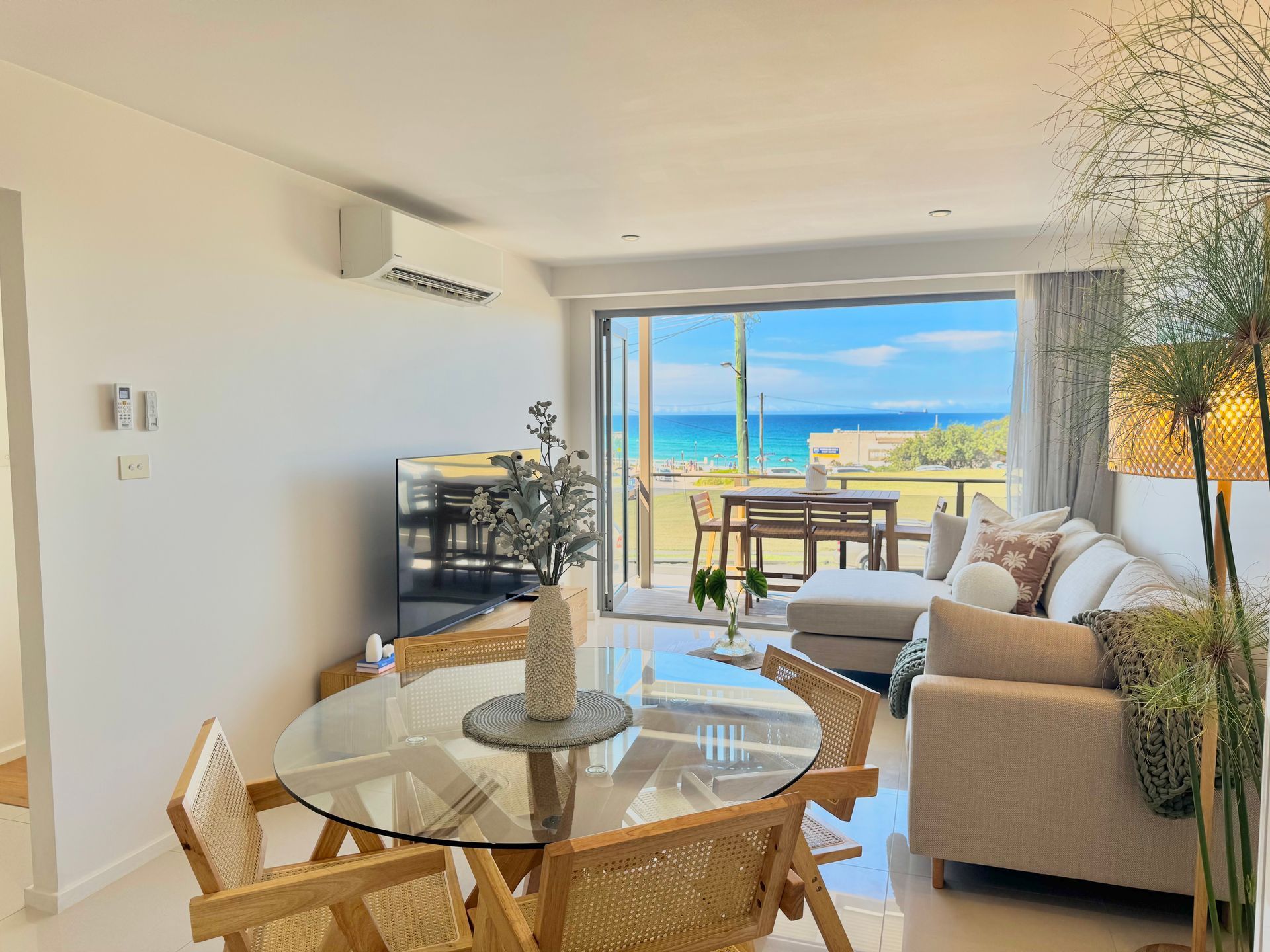 A sunlit living and dining area with a glass table, neutral sofa, and large sliding glass doors overlooking the ocean.