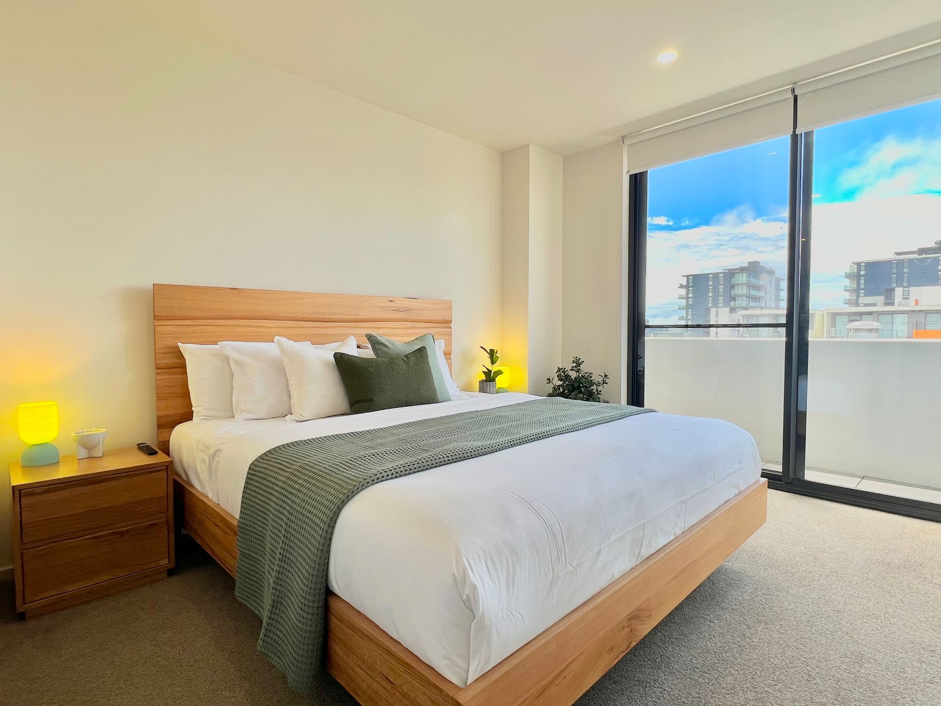 A modern bedroom with a wooden bed frame, white bedding, green accents, a matching nightstand, and large glass windows.