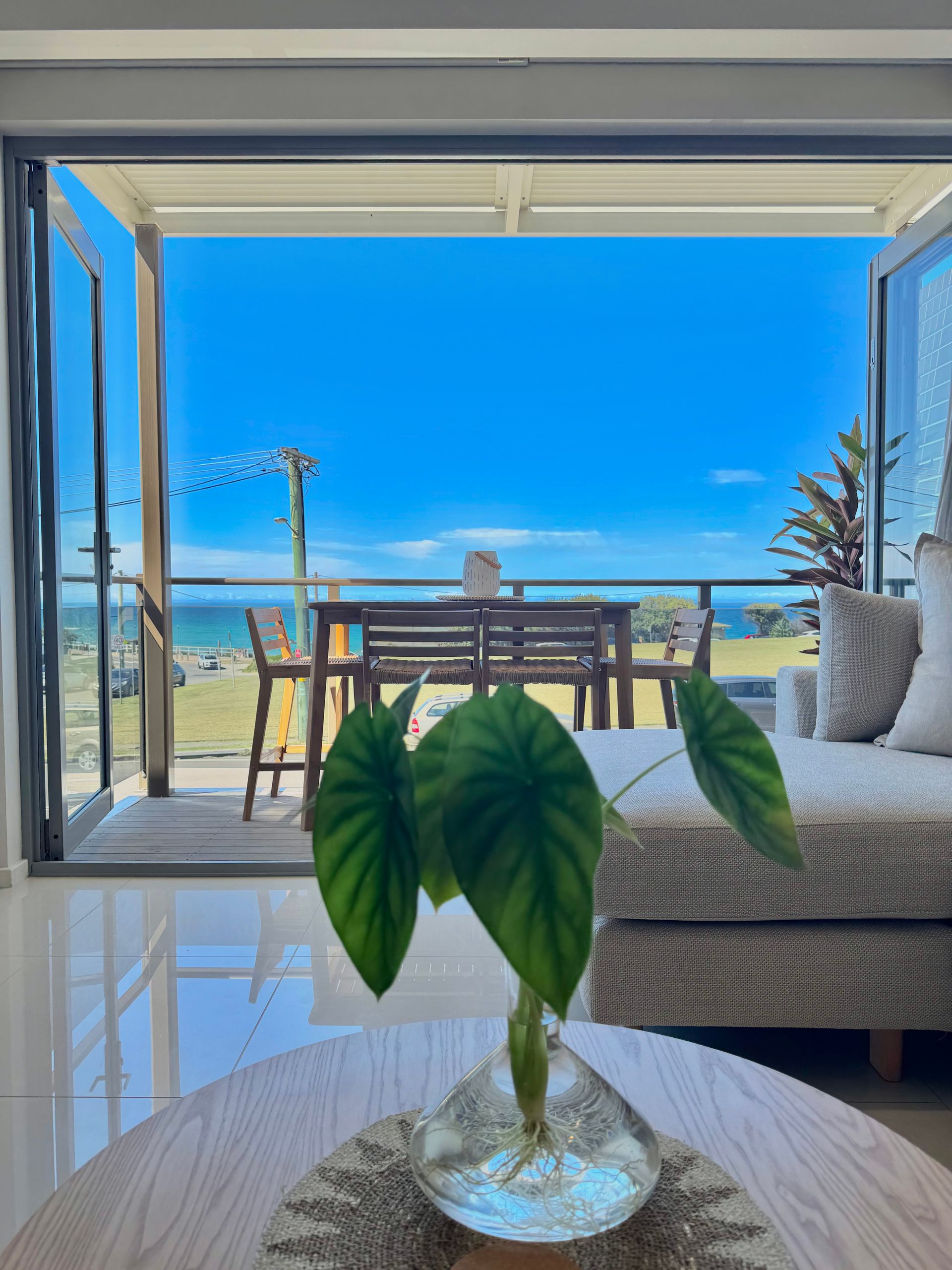 A small plant in a glass vase sits on a round table, overlooking an ocean view from a modern, open-air living space.