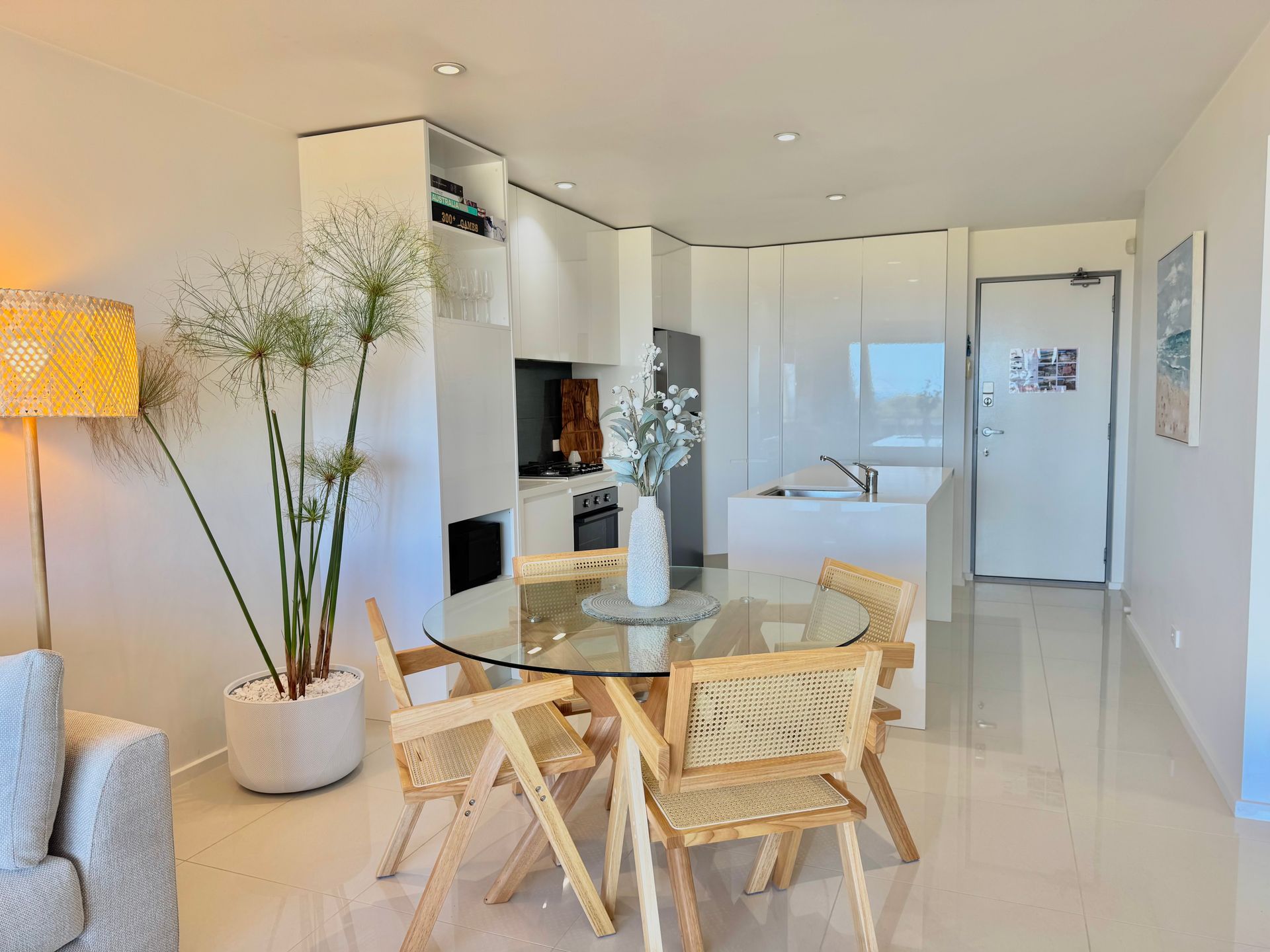 A modern dining area with a circular glass table, four wooden chairs, a large potted plant, and a minimalist kitchen.