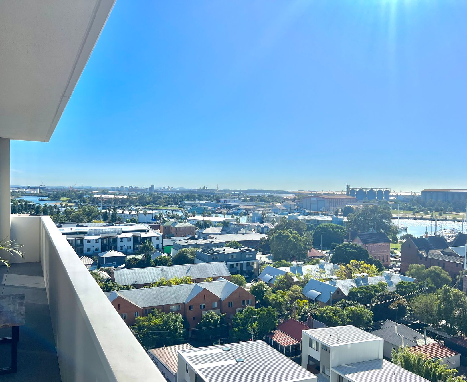 A high-angle view from a balcony overlooking a city neighborhood, rooftops, and distant blue water under a clear sky.