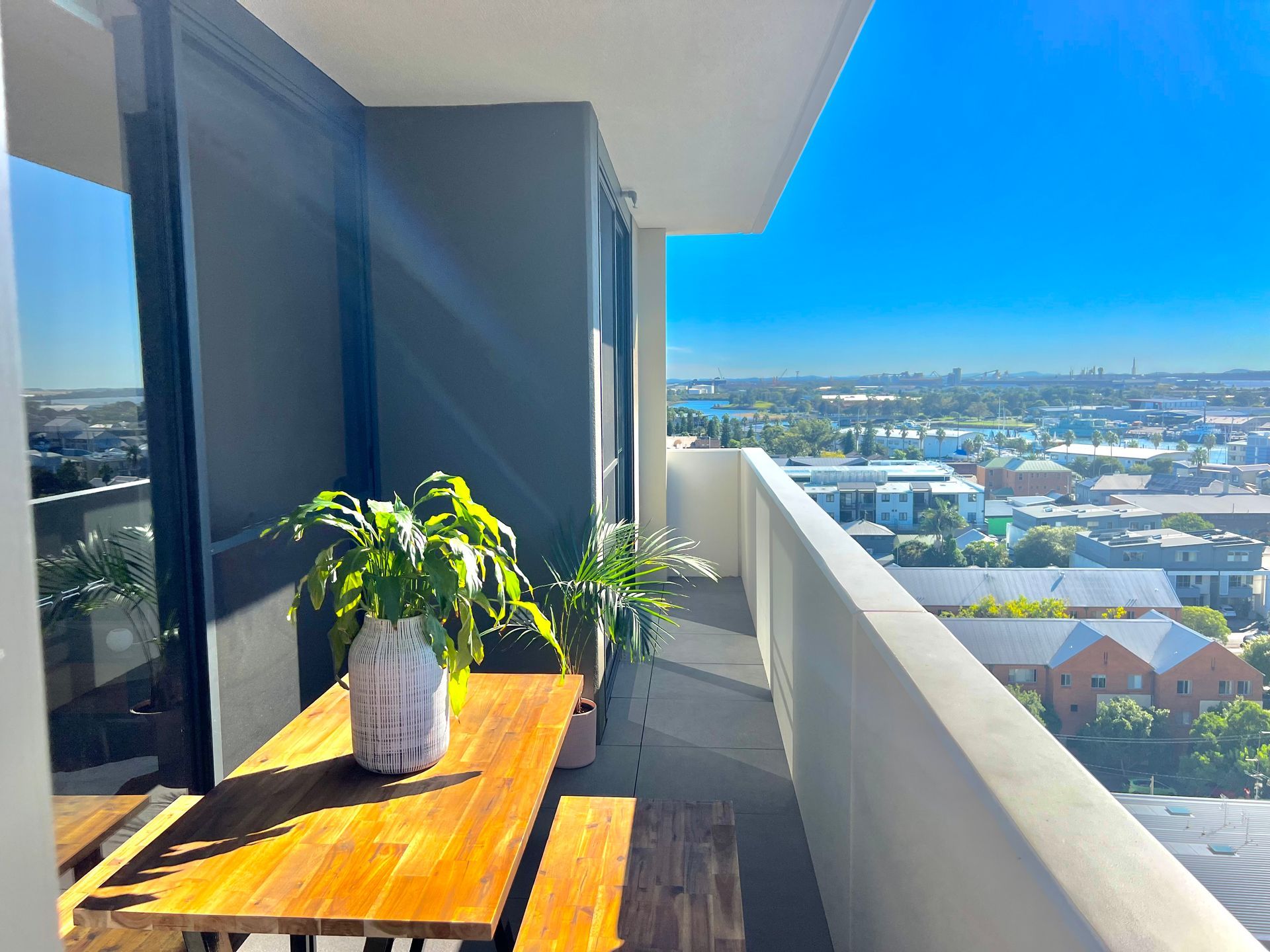 A balcony with a wooden table and bench, a potted plant, and a wide view of a suburban landscape under a clear blue sky.