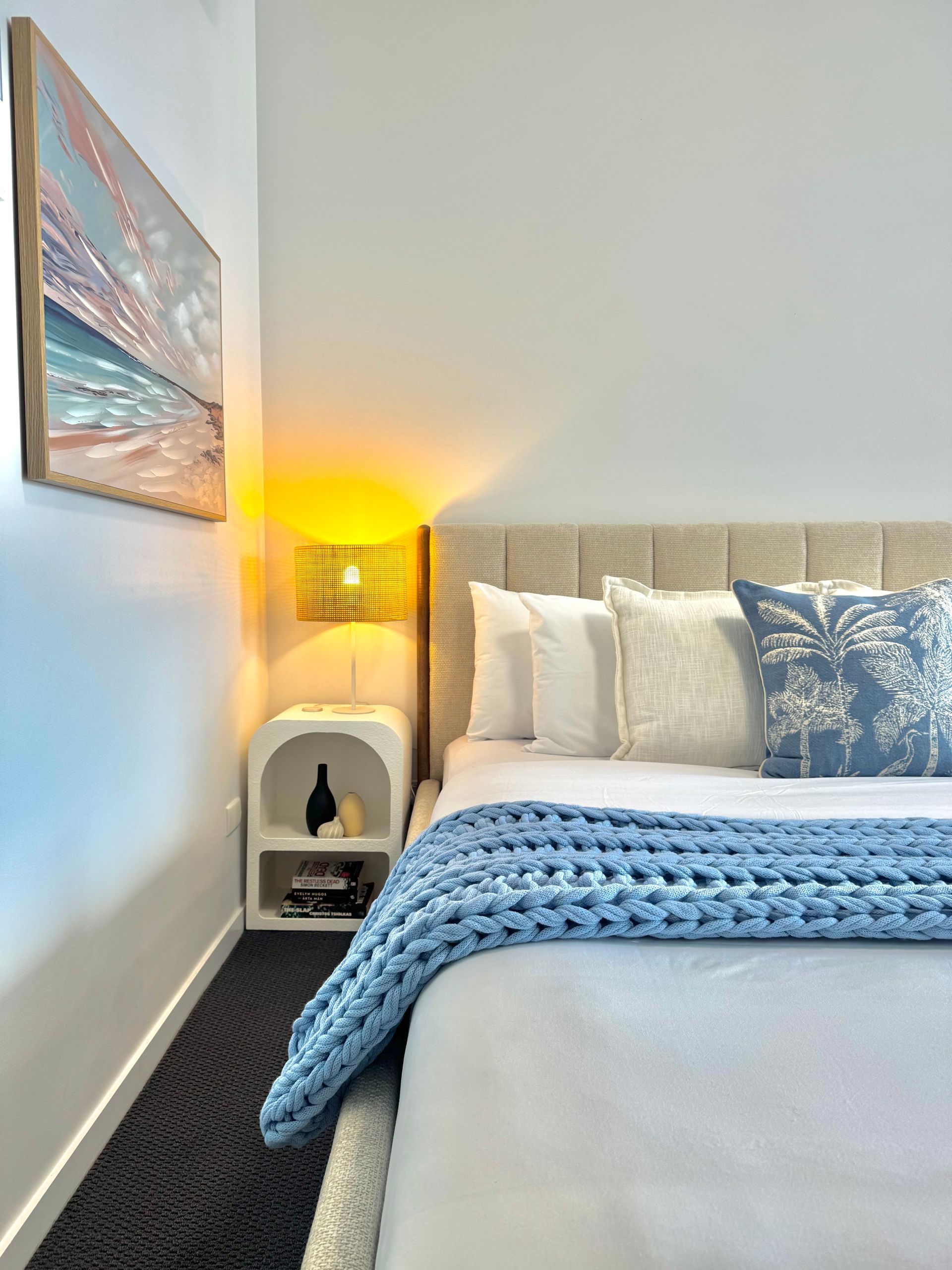 A bedroom with a textured cream headboard, blue and white pillows, a chunky blue throw blanket, and a glowing bedside lamp.