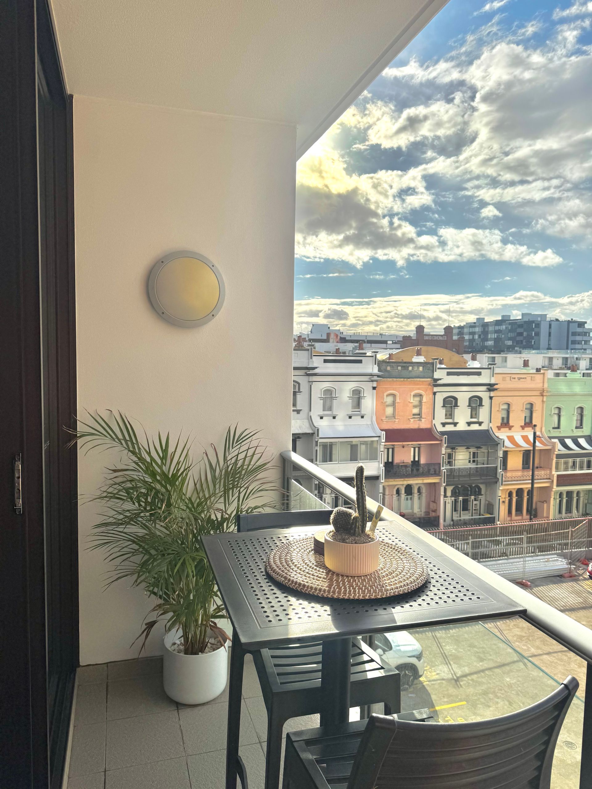 A balcony overlooking a city street with a small table, two chairs, and a potted plant against a white wall at sunset.