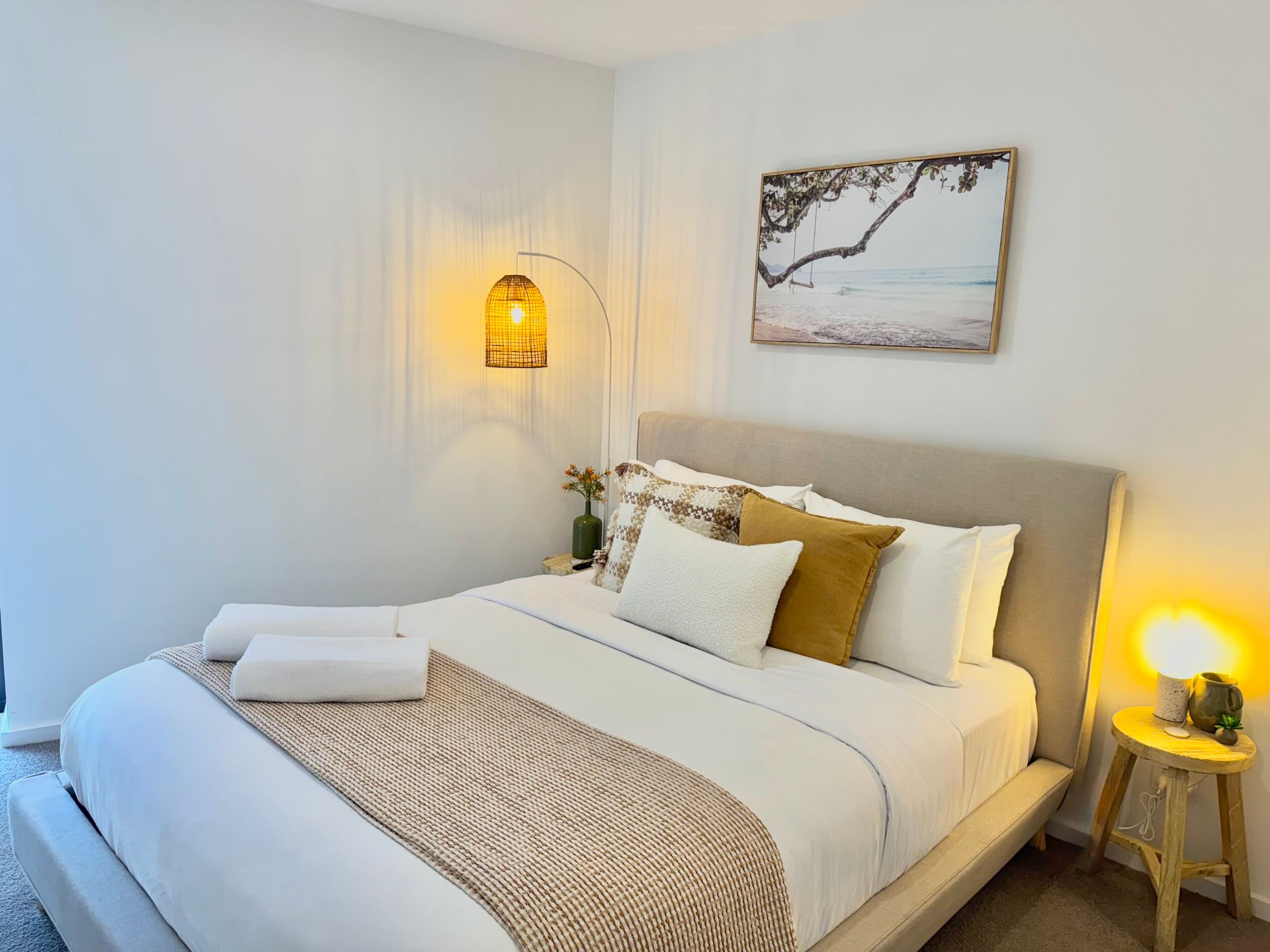 A cozy bedroom with a neutral-toned bed, a woven hanging lamp, a landscape wall art piece, and a small bedside stool.