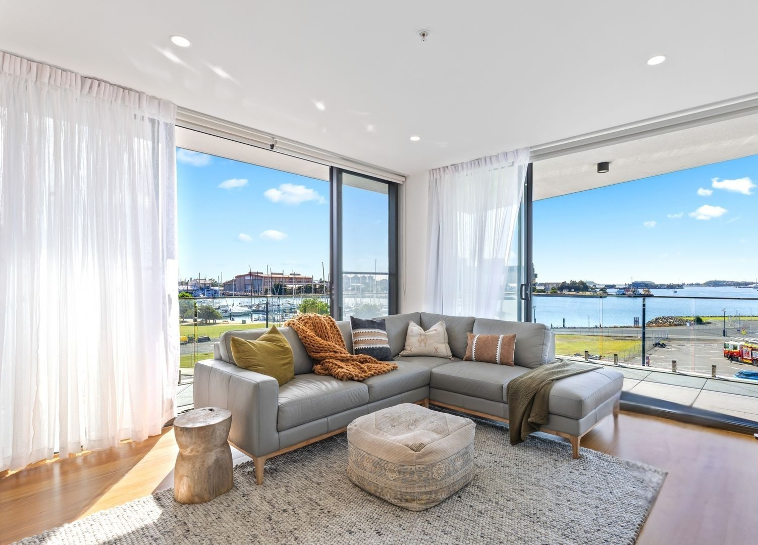 A grey L-shaped sofa with pillows sits on a light rug in a sunlit living room with floor-to-ceiling windows viewing water.