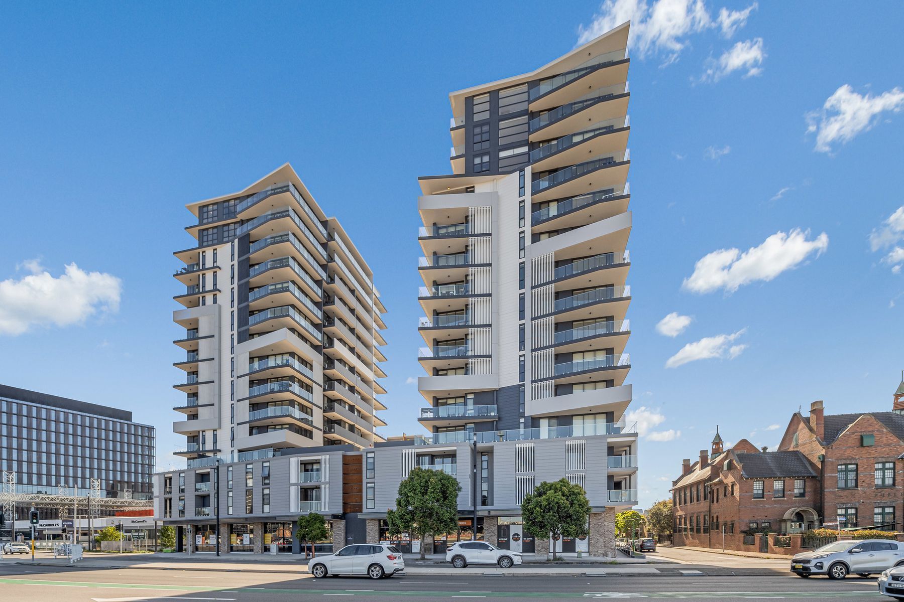 Two modern high-rise apartment towers with balconies under a bright blue sky, with older brick buildings to the right.