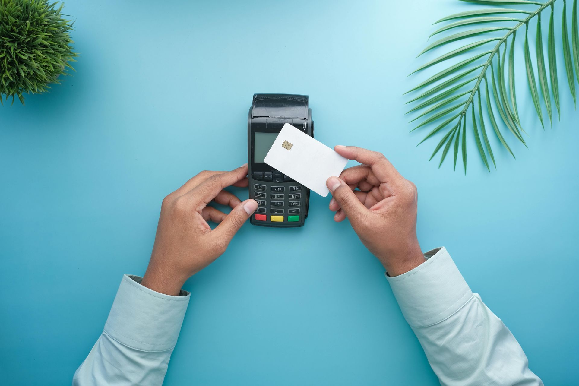Hands holding a credit card over a payment terminal on a blue surface.