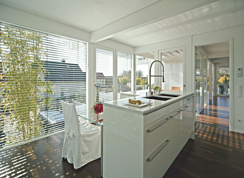 Moderne weiße Küche mit Kochinsel, großen Fenstern mit Jalousien, Blick auf Bäume und Häuser.