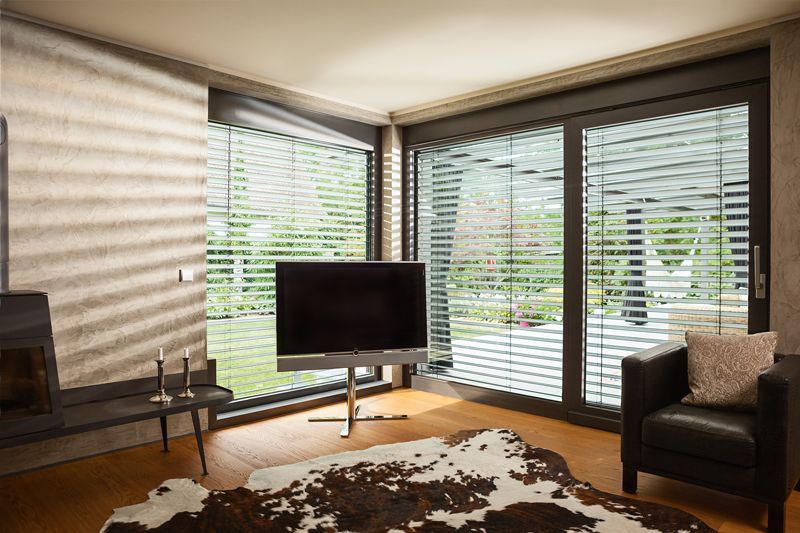 Wohnzimmer mit großen Fenstern, Jalousien, Fernseher, Ledersessel und Kuhfellteppich.