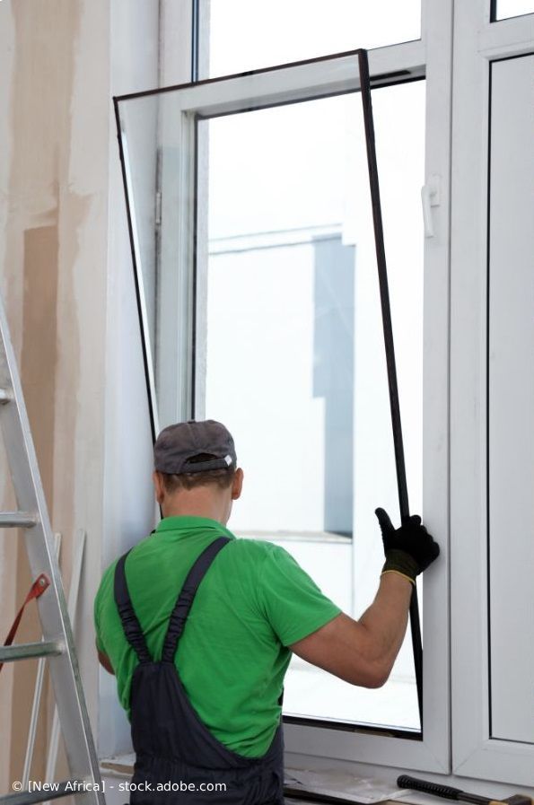 Person, die ein Fliegengitter montiert; weißer Fensterrahmen, grünes Hemd, schwarze Handschuhe.