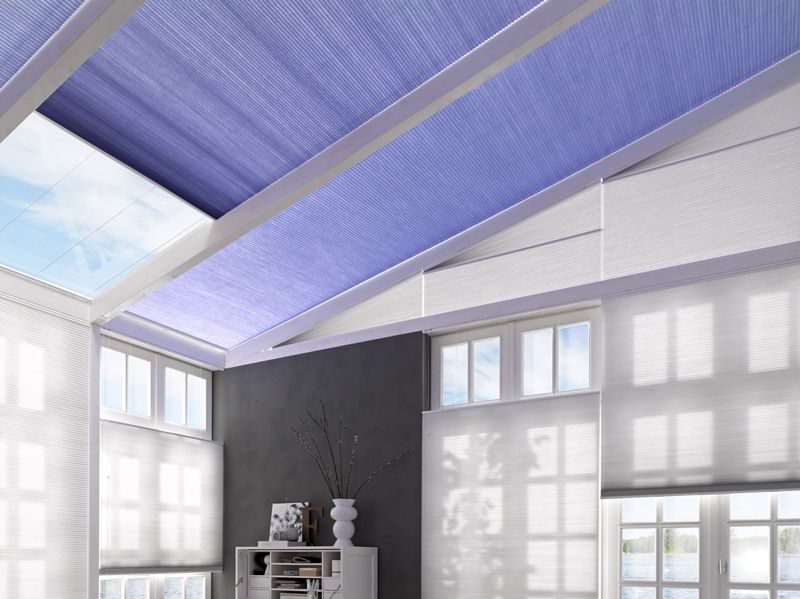 Blaue und weiße Stoffrollos verdecken ein Dachfenster und die Fenster in einem hellen Raum.