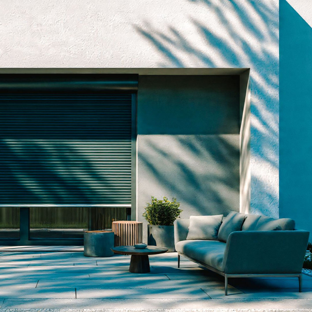 Terrasse extérieure avec canapé turquoise, plantes et volets verts fermés ; ombres projetées par les feuilles de palmier.