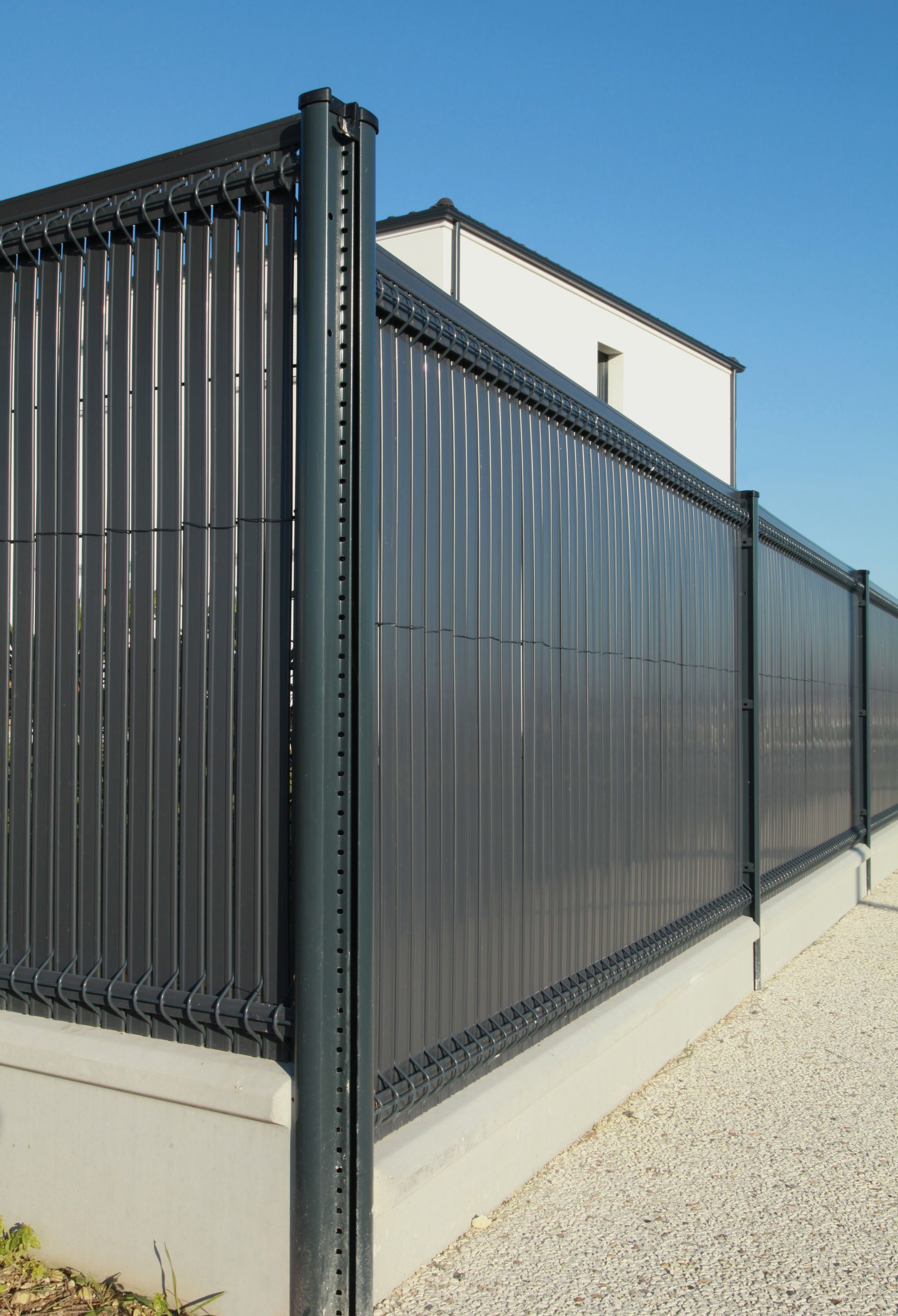 Clôture à lattes verticales gris foncé délimitant une zone de gravier, avec un bâtiment blanc en arrière-plan.