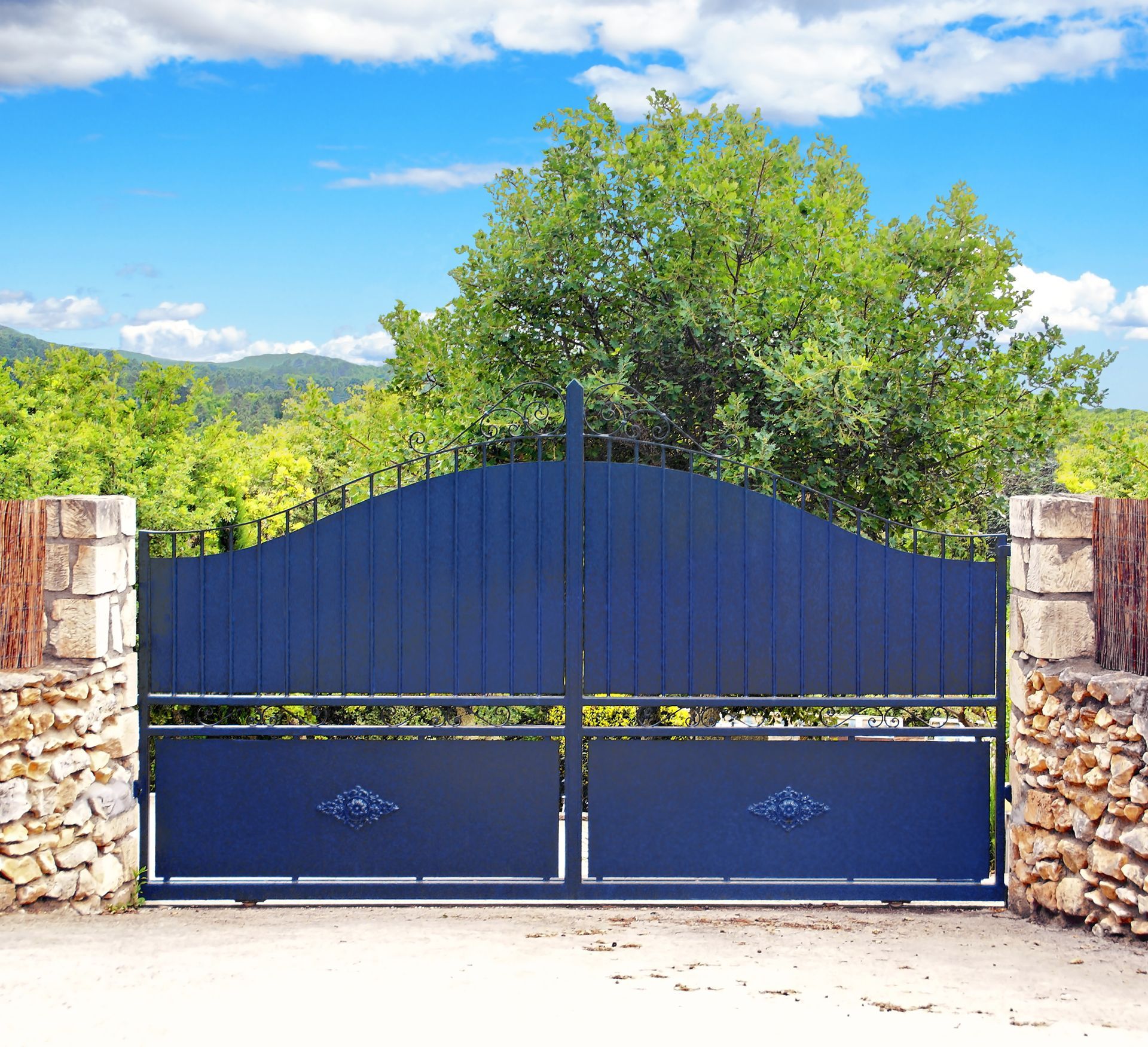 Portail en métal bleu, piliers de pierre et feuillage vert sur fond de ciel bleu.