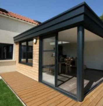 Véranda avec boiseries gris foncé, grandes portes vitrées et terrasse en bois attenante à une maison en briques claires.