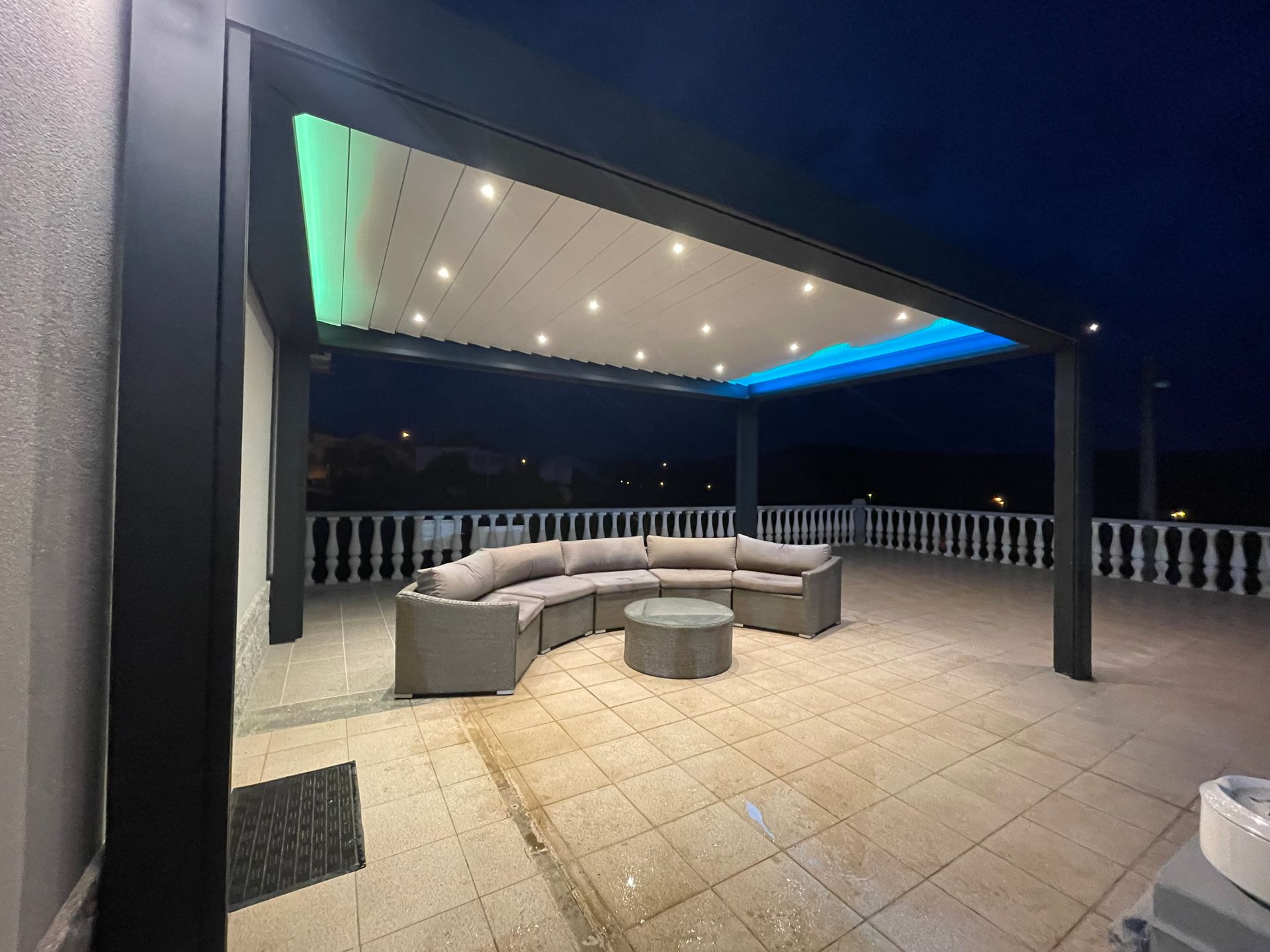 Terrasse avec mobilier gris sous une pergola sombre illuminée de lumières colorées la nuit.