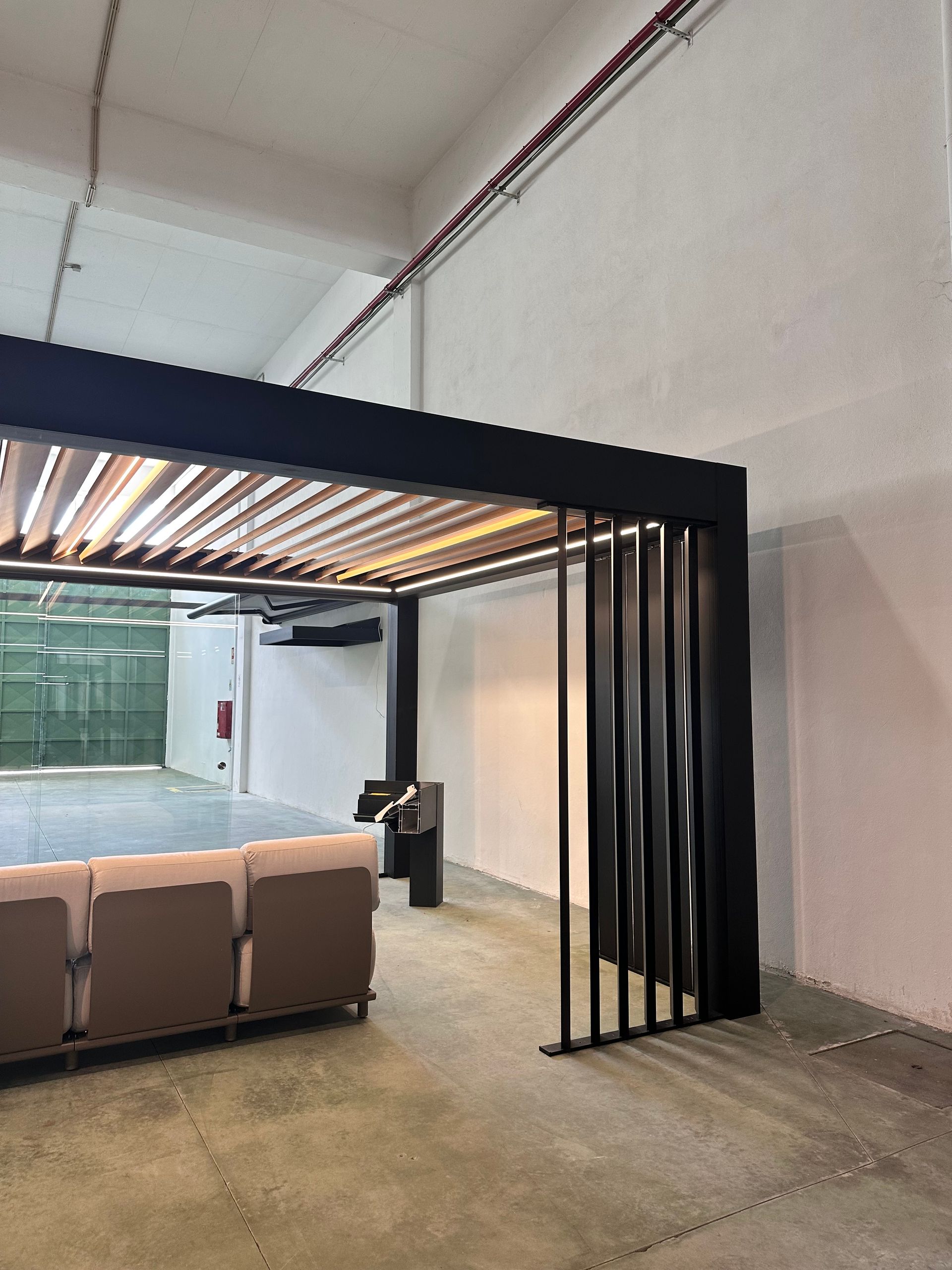 Pergola noire à toit à lattes et lattes verticales noires, au-dessus d'un canapé dans un intérieur aux murs blancs.