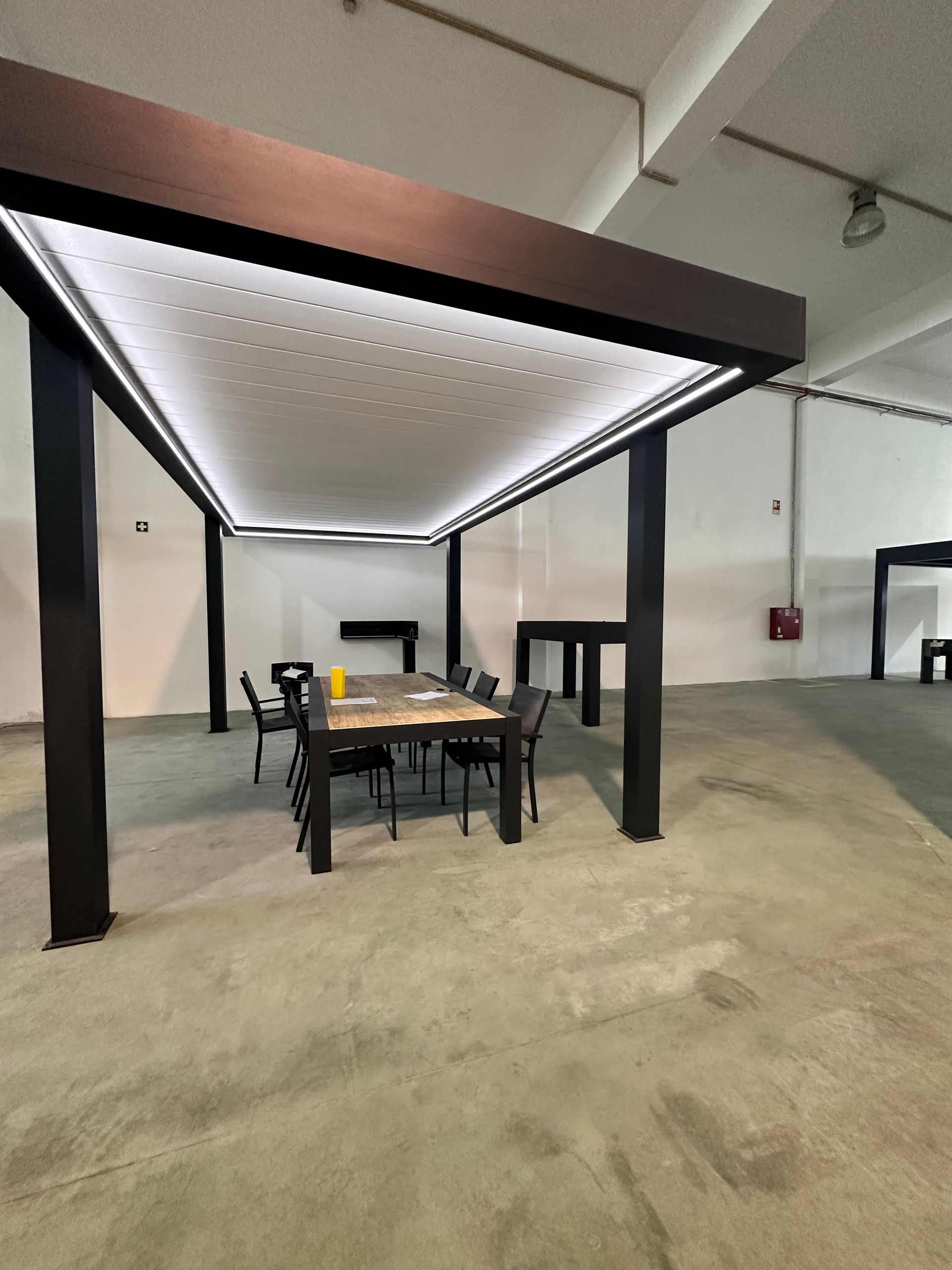 Pergola noire avec un toit blanc perforé, table et chaises dans un espace au sol en béton.