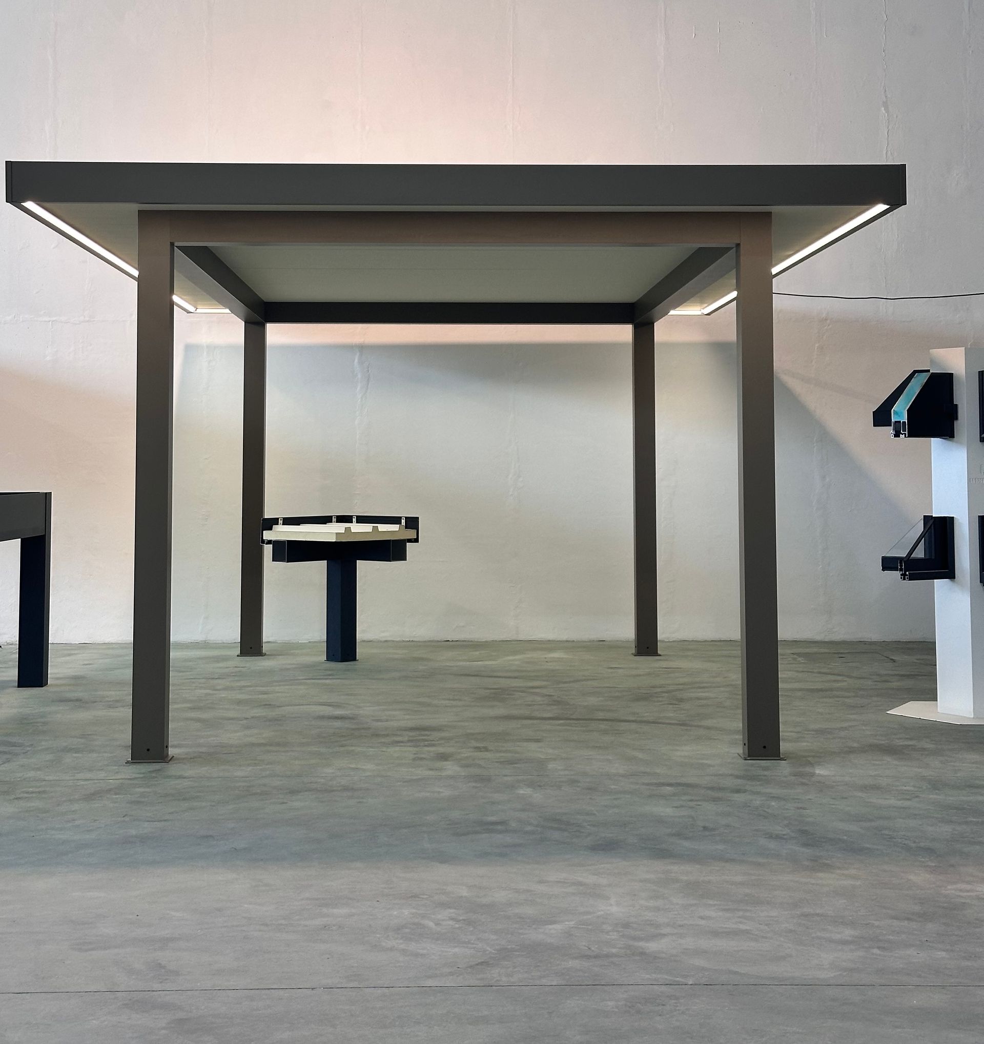 Pergola rectangulaire grise avec éclairage intégré, dans une pièce au sol gris, avec une autre table en arrière-plan.