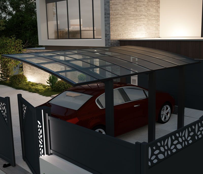 Une voiture rouge garée sous un abri à toit transparent, à côté d'une maison et derrière une clôture décorative.