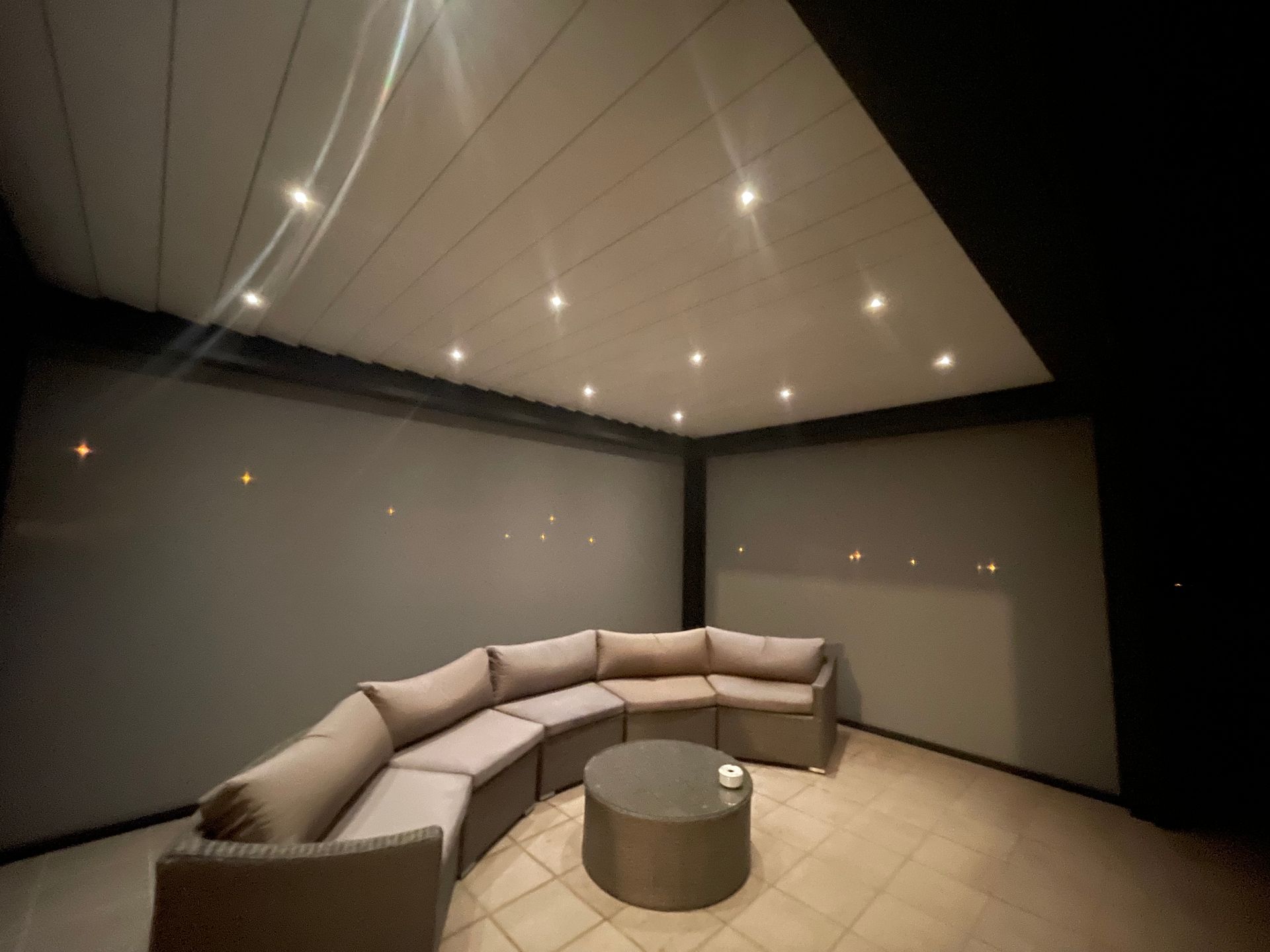 Canapé d'angle dans une pièce extérieure sombre avec spots encastrés au plafond et table basse ronde.