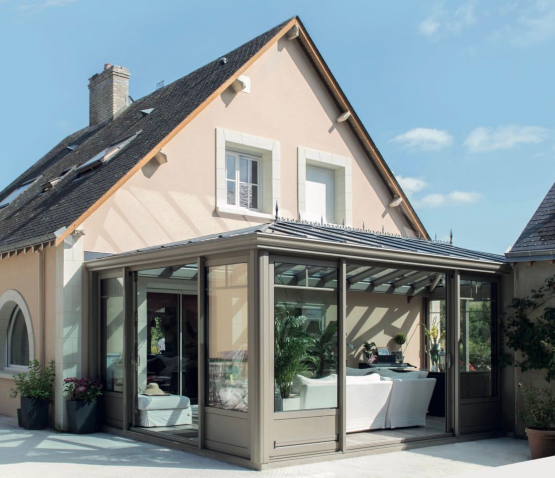 Véranda beige attenante à une maison rose pâle. Plantes vertes à l'intérieur, ciel bleu.