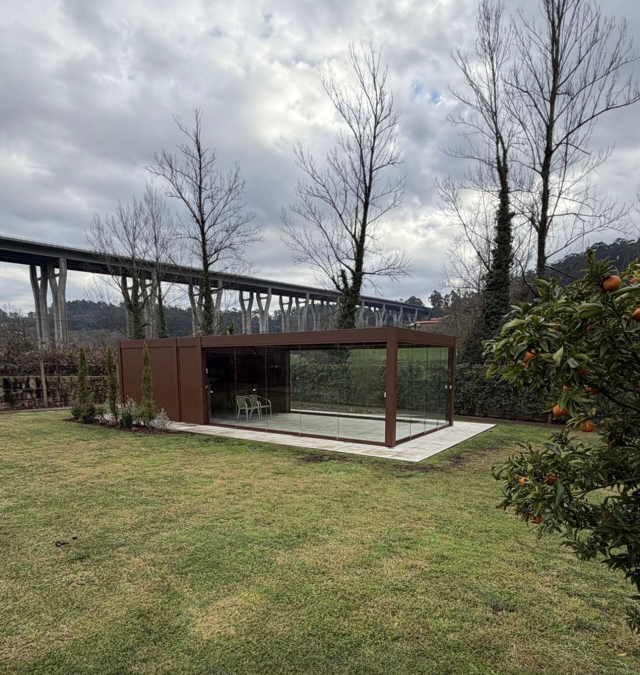 Pool house marron aux parois de verre, avec un pont en arrière-plan, situé dans un jardin gazonné.