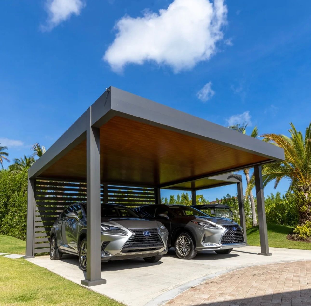 Deux voitures garées sous un abri moderne gris avec un plafond en bois, sur fond de ciel bleu.