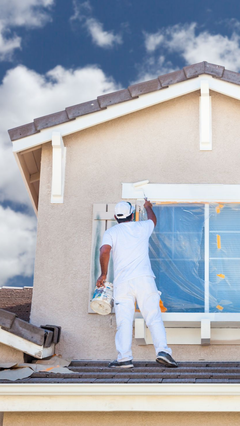 Une personne en salopette blanche peint les encadrements de fenêtres sur la façade d'une maison.