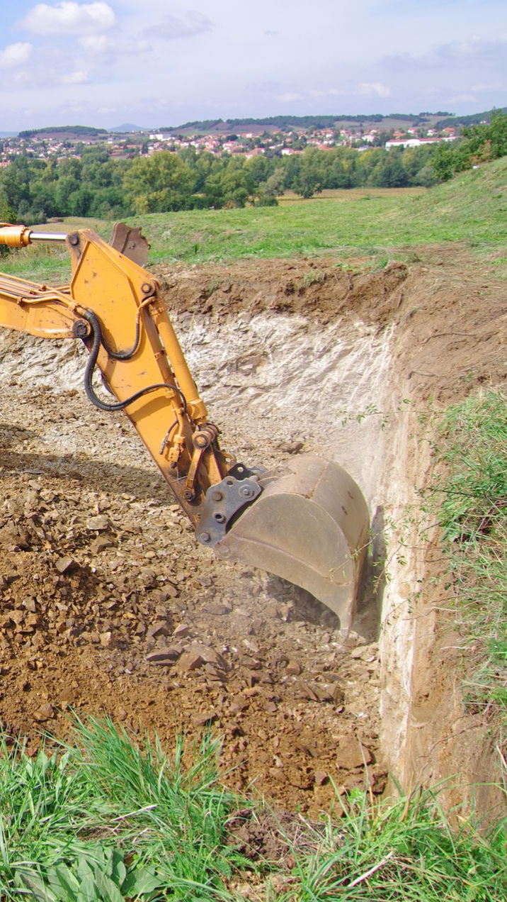 Une pelleteuse jaune creuse une tranchée dans la terre, avec un paysage urbain au loin.