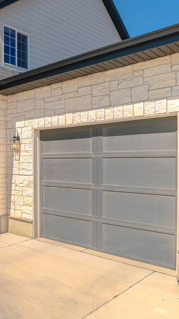 Porte de garage grise sur une maison avec un revêtement en pierre claire et une fenêtre au-dessus.