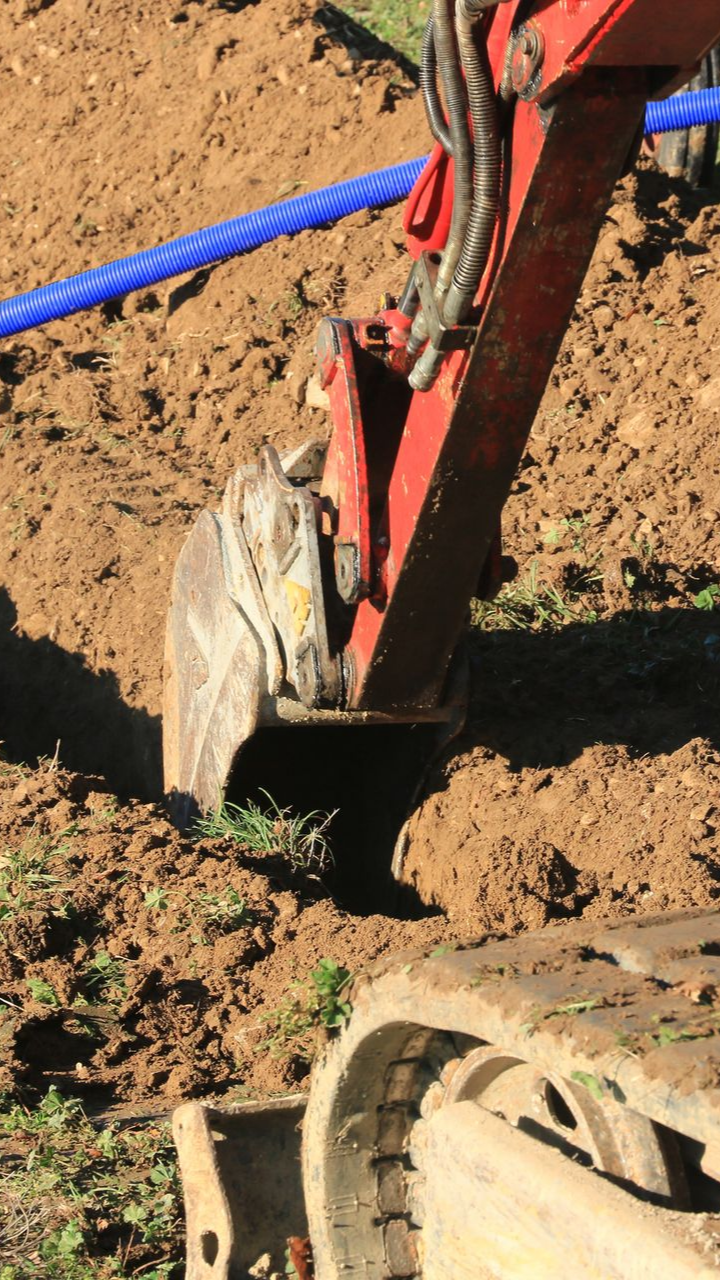 Godet d'excavatrice rouge creusant une tranchée dans un sol brun, avec un tuyau bleu visible au-dessus.